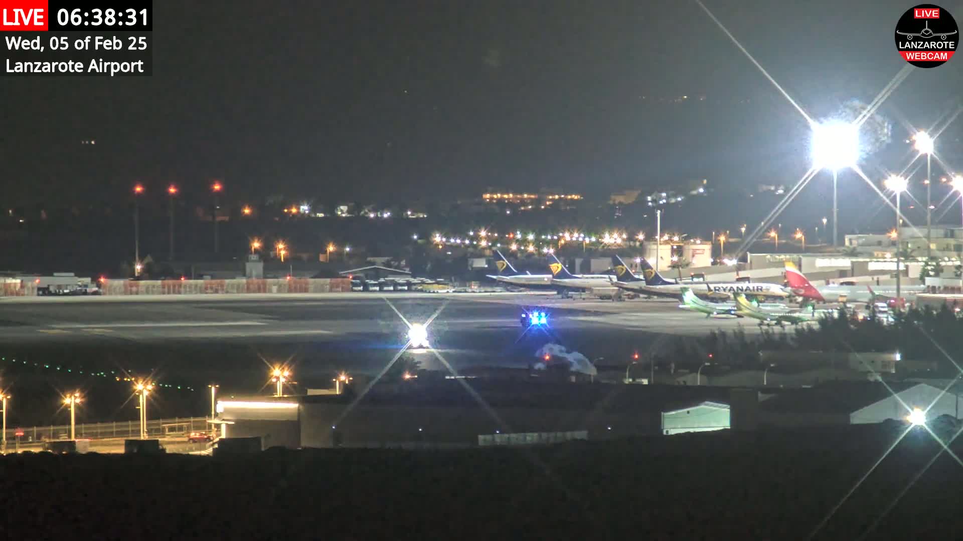 A nighttime view of an airport tarmac with several airplanes parked, bright lights illuminating the scene, and a vehicle with flashing blue lights.