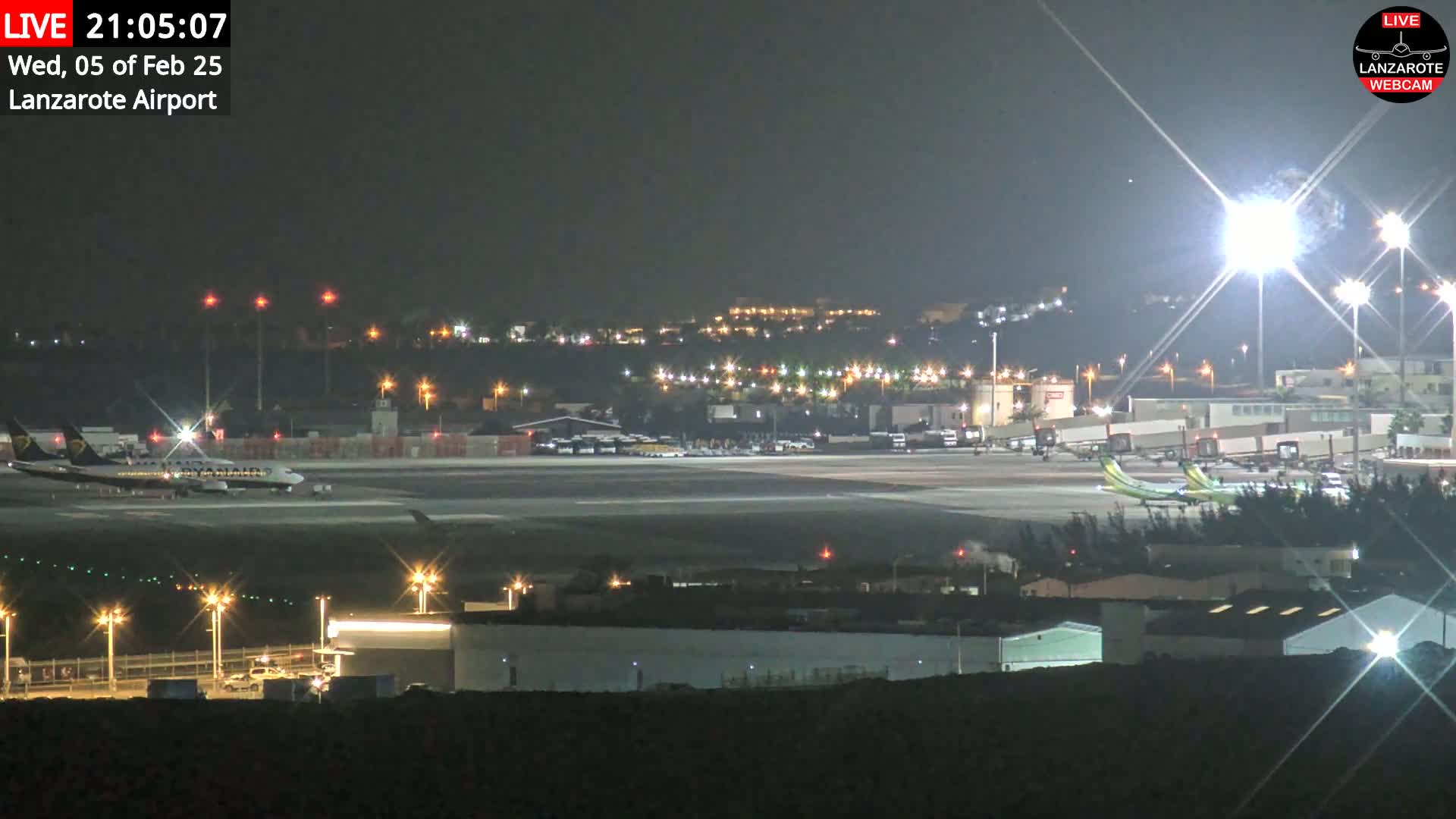 A nighttime view of an airport tarmac with several airplanes, brightly lit buildings, and a city skyline in the background under clear skies.