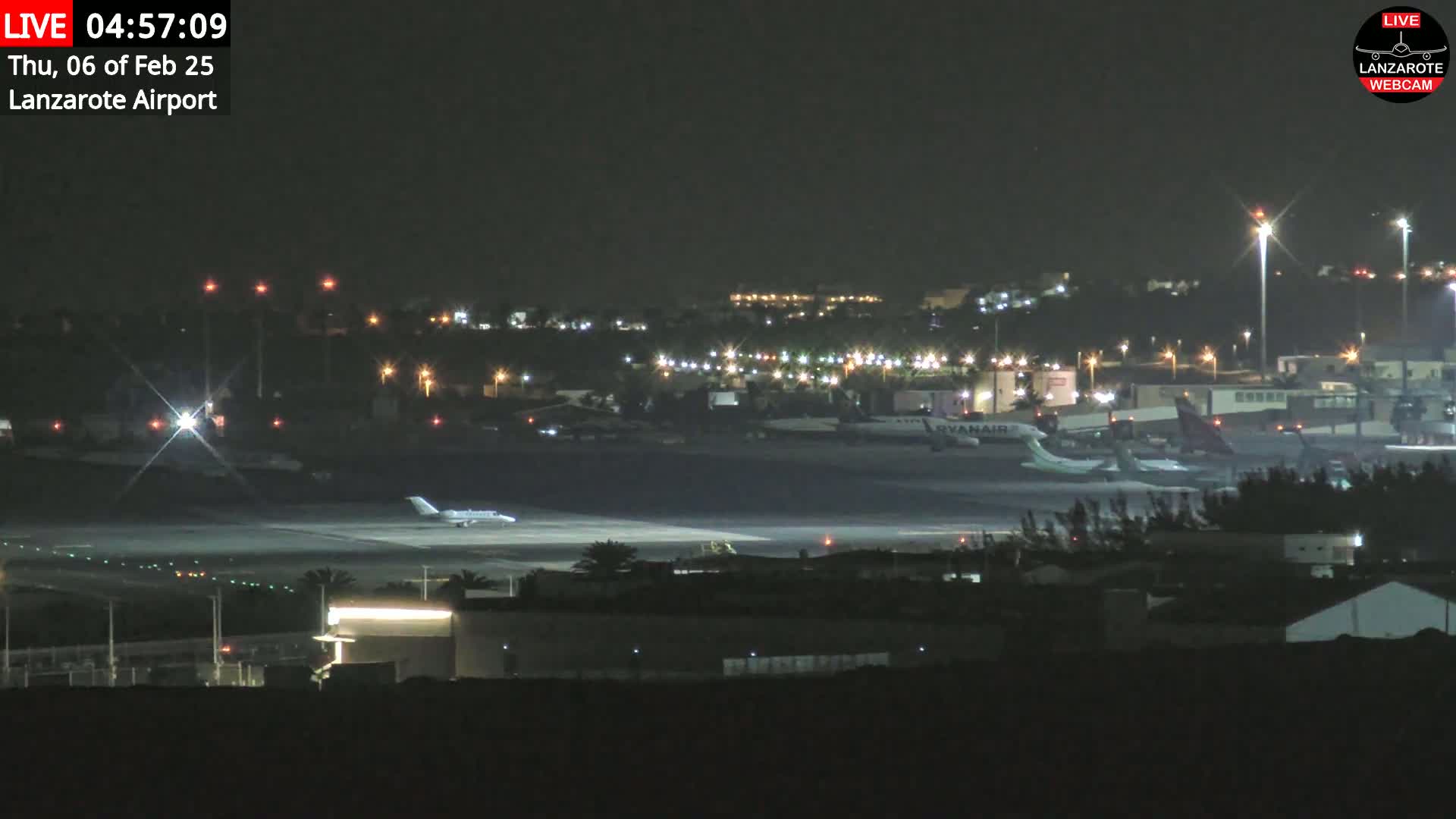 A nighttime view of an airport runway with several airplanes, including one taxiing, and surrounding buildings illuminated by lights.