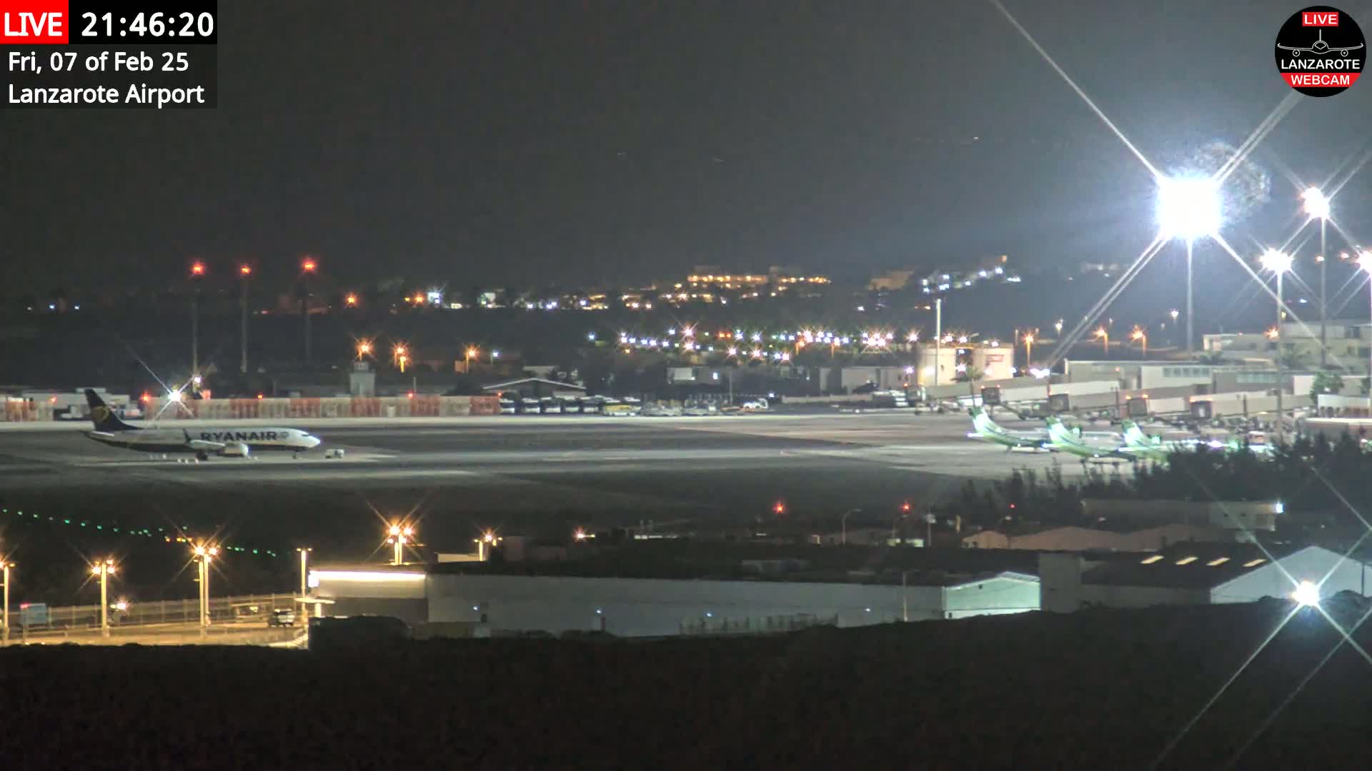 At night, a Ryanair airplane is parked on a runway at an airport, with other planes visible in the distance under bright lights.
