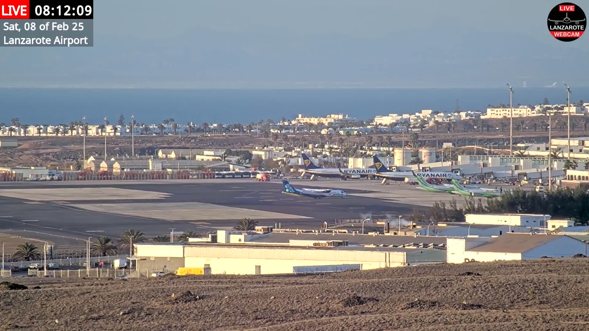 An airport tarmac with several airplanes, including two Ryanair jets, is visible, with a calm ocean and coastal buildings in the background under a clear, bright sky.