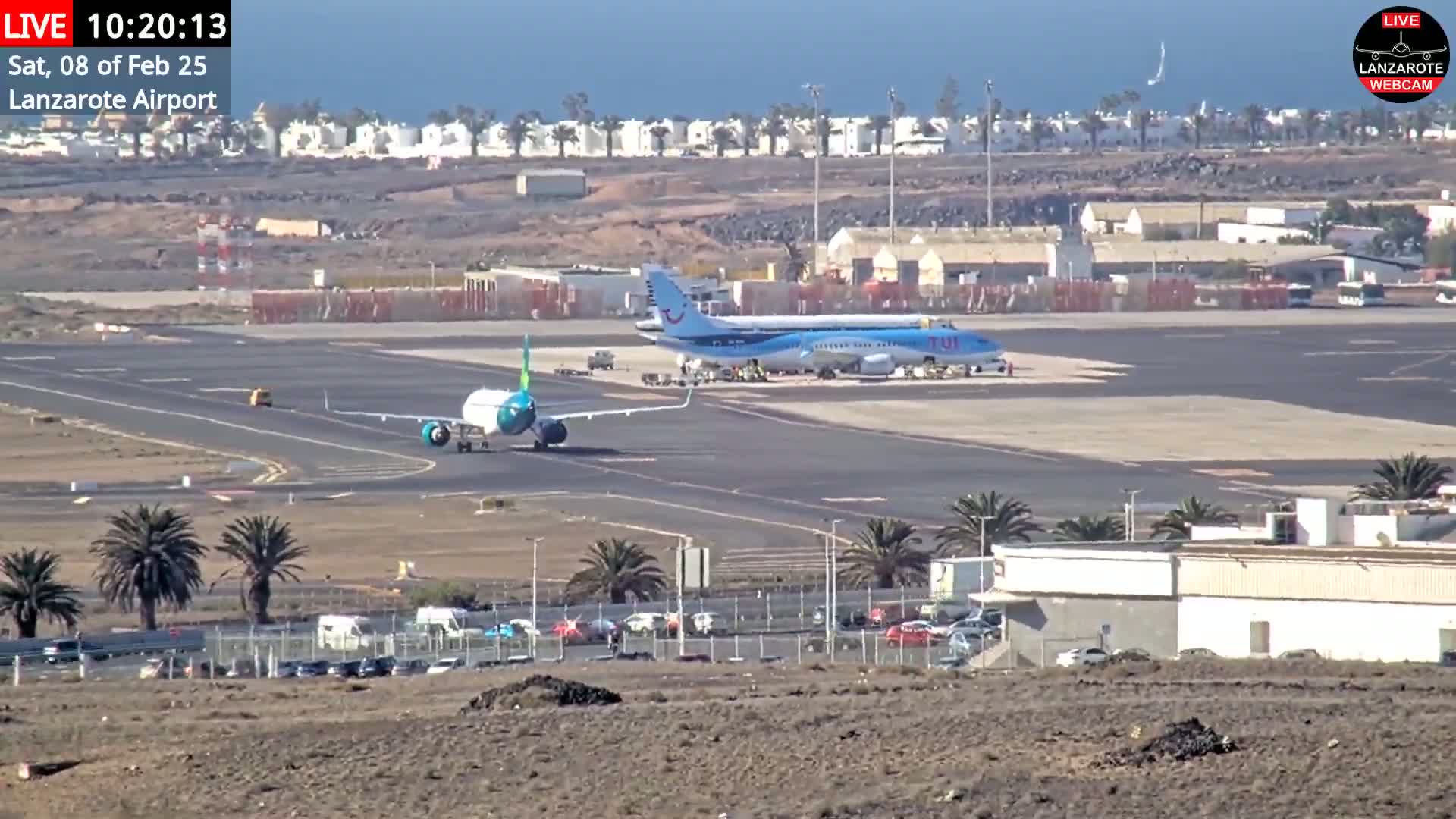 Two airplanes are on an airport tarmac under a mostly sunny sky, with palm trees and buildings visible in the background.