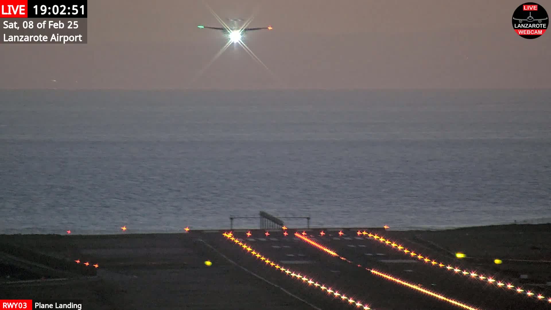 A plane is landing on a runway at dusk, with the ocean visible in the background.
