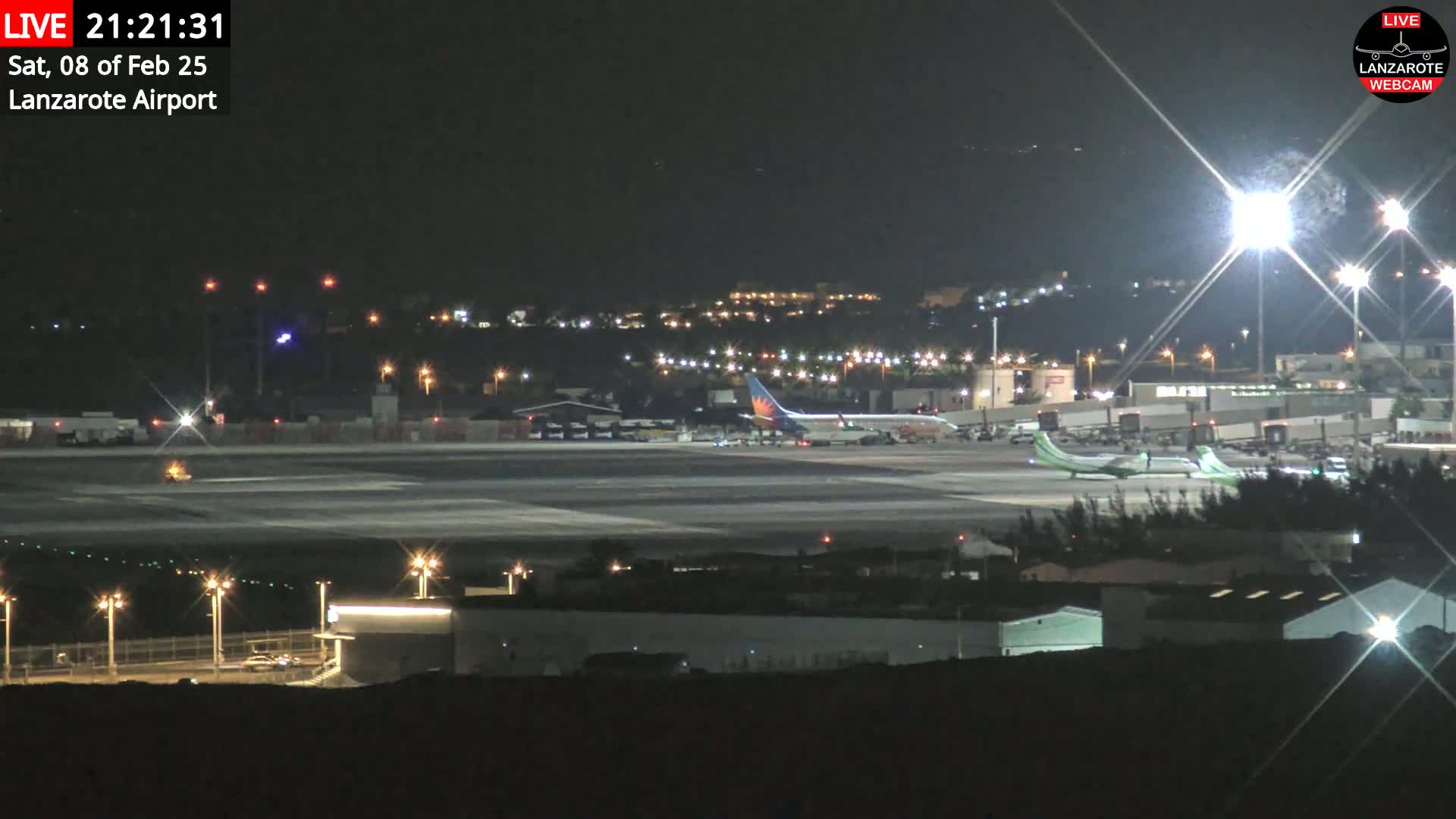 A nighttime, brightly lit airport tarmac with several airplanes parked and one on the runway.