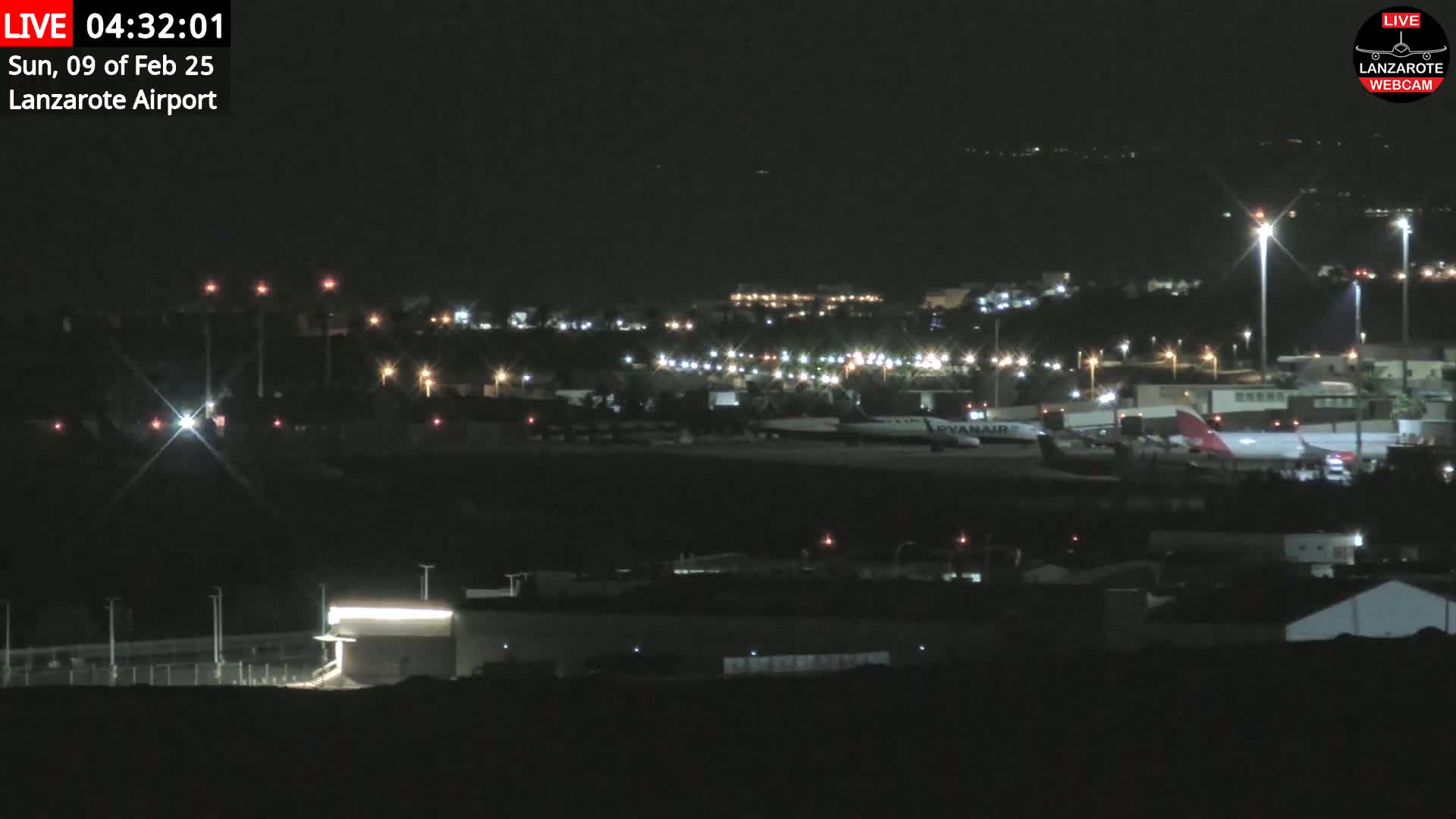 A nighttime long shot of an airport tarmac with several airplanes, surrounded by dark buildings and illuminated by lights.
