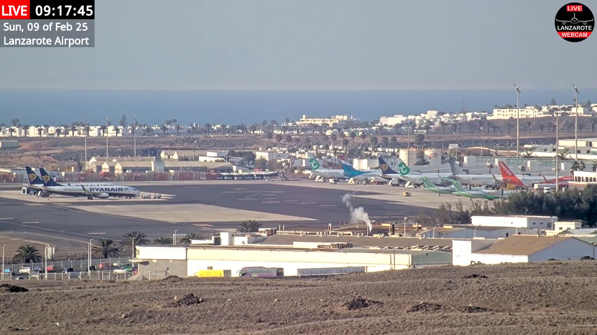 A long-range view of an airport tarmac with several airplanes parked, surrounded by buildings and a coastal landscape under clear skies.