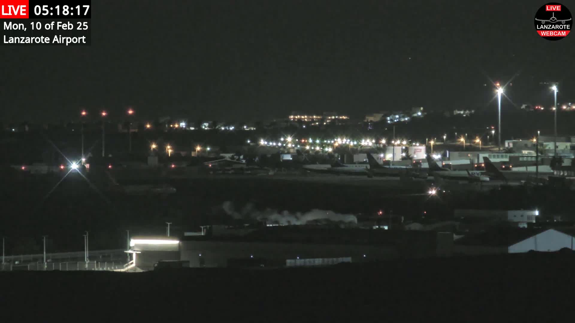 A nighttime view of an airport tarmac with several airplanes parked, illuminated by lights, and some steam or smoke visible in the foreground.