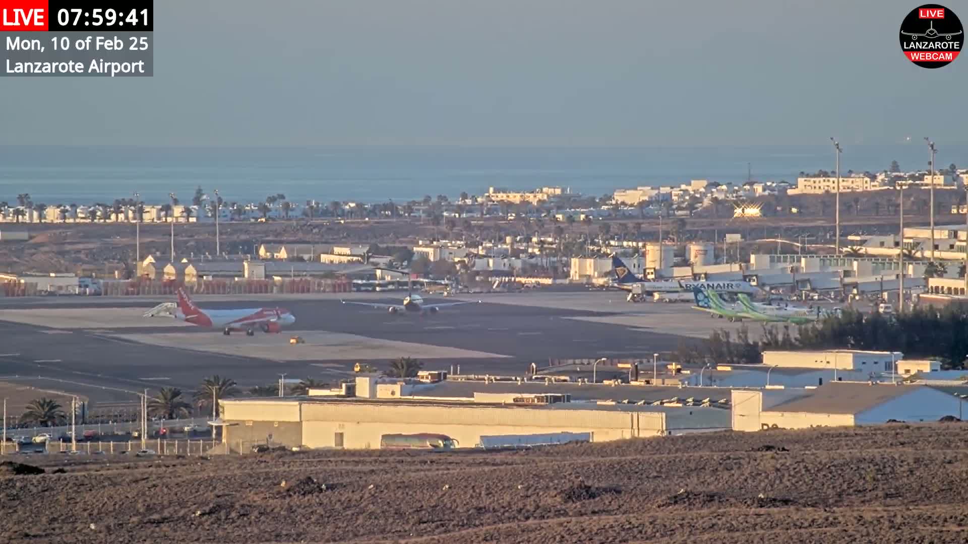 A calm, sunny morning view of an airport tarmac with several airplanes, including one being serviced, and a coastal town visible in the background.