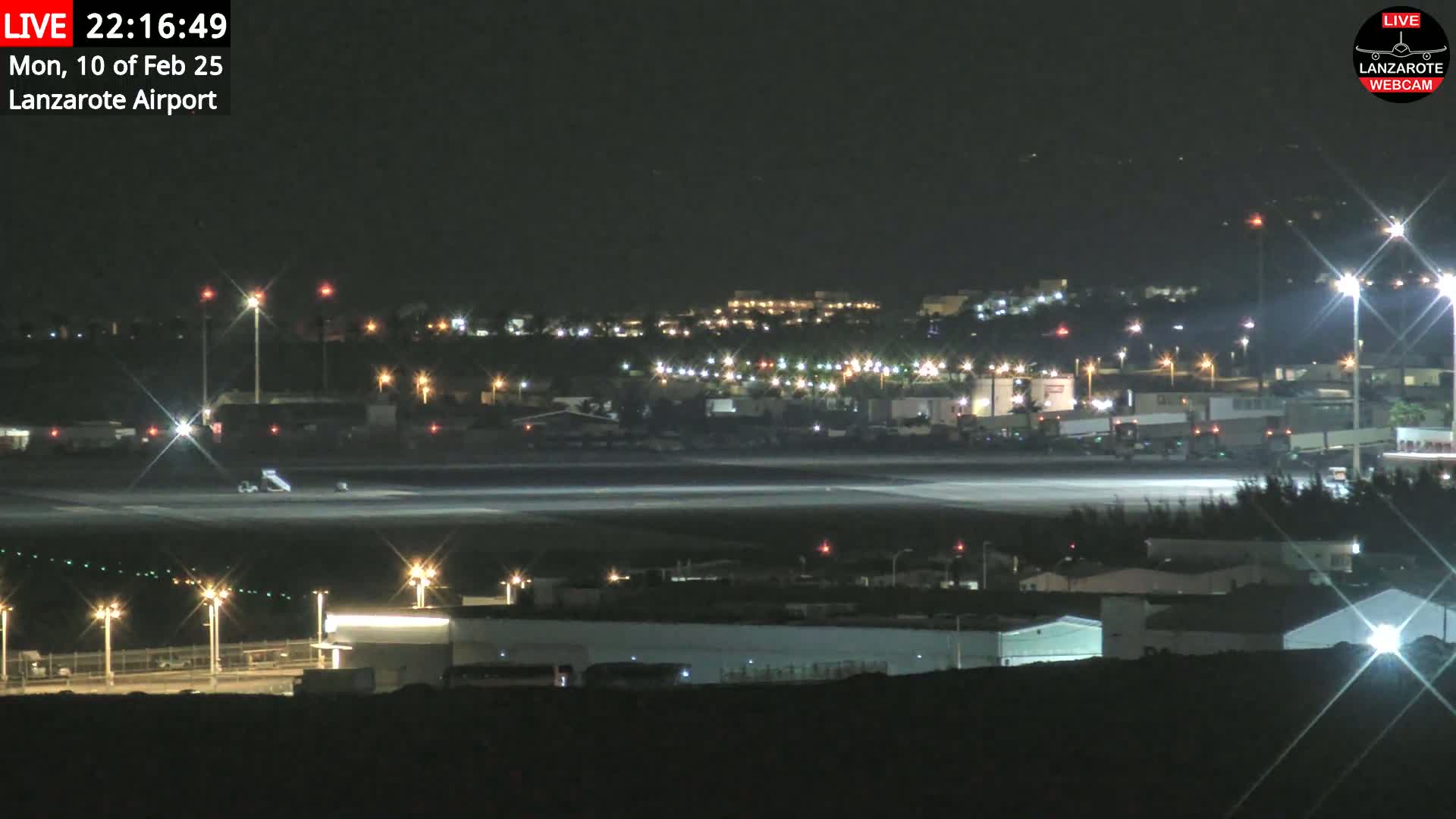 A nighttime view of an airport runway, with airport buildings and lights illuminated, under a clear sky.