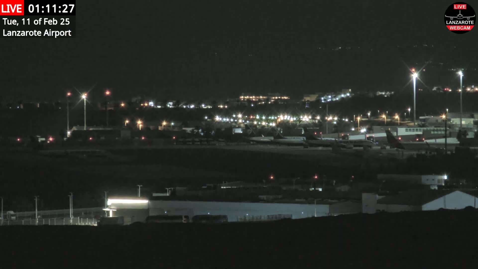 A nighttime, dimly lit view of an airport tarmac with several airplanes and surrounding buildings under a clear sky.