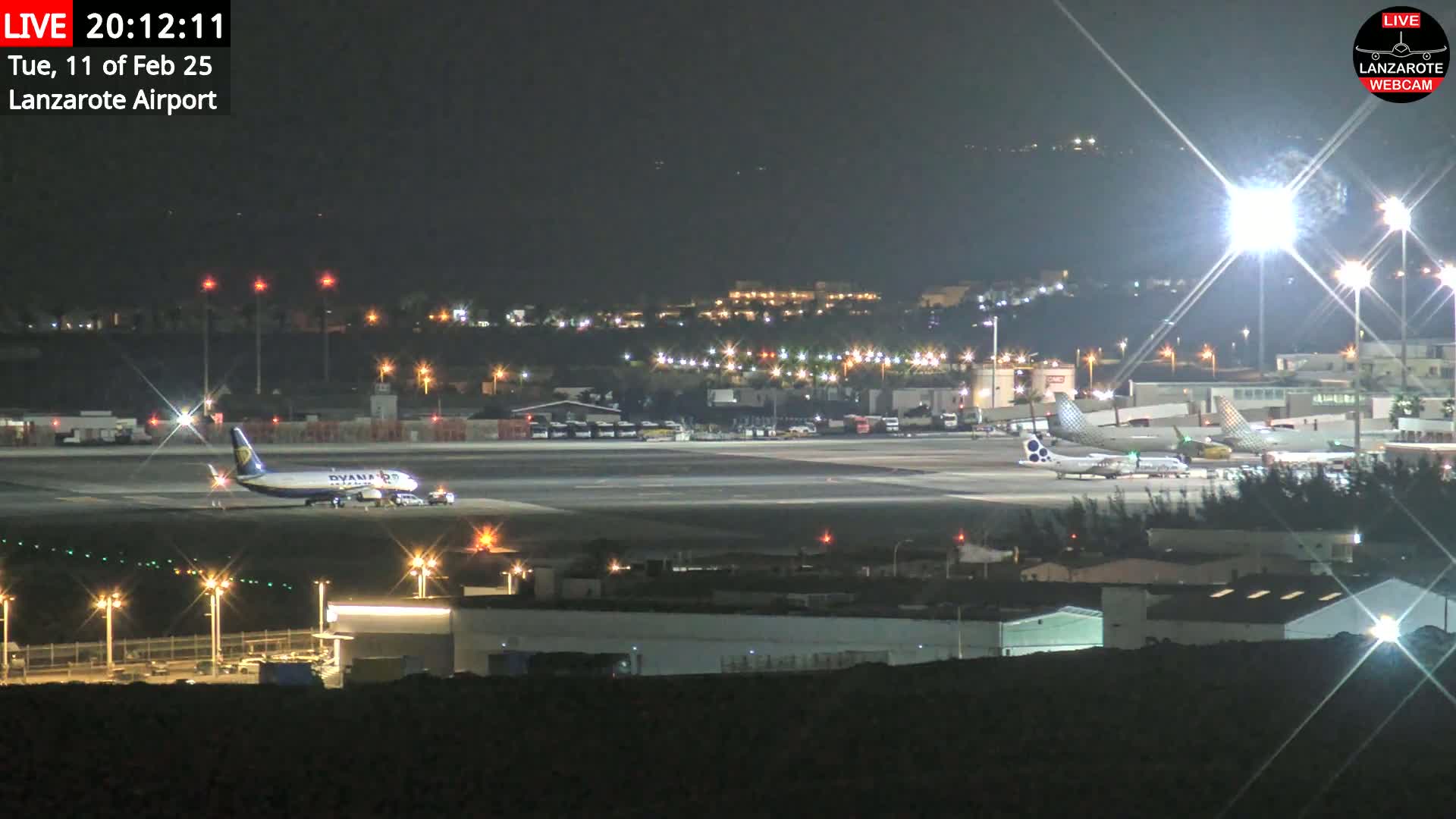A nighttime view of an airport tarmac with several airplanes, brightly lit buildings, and ground support vehicles under clear skies.