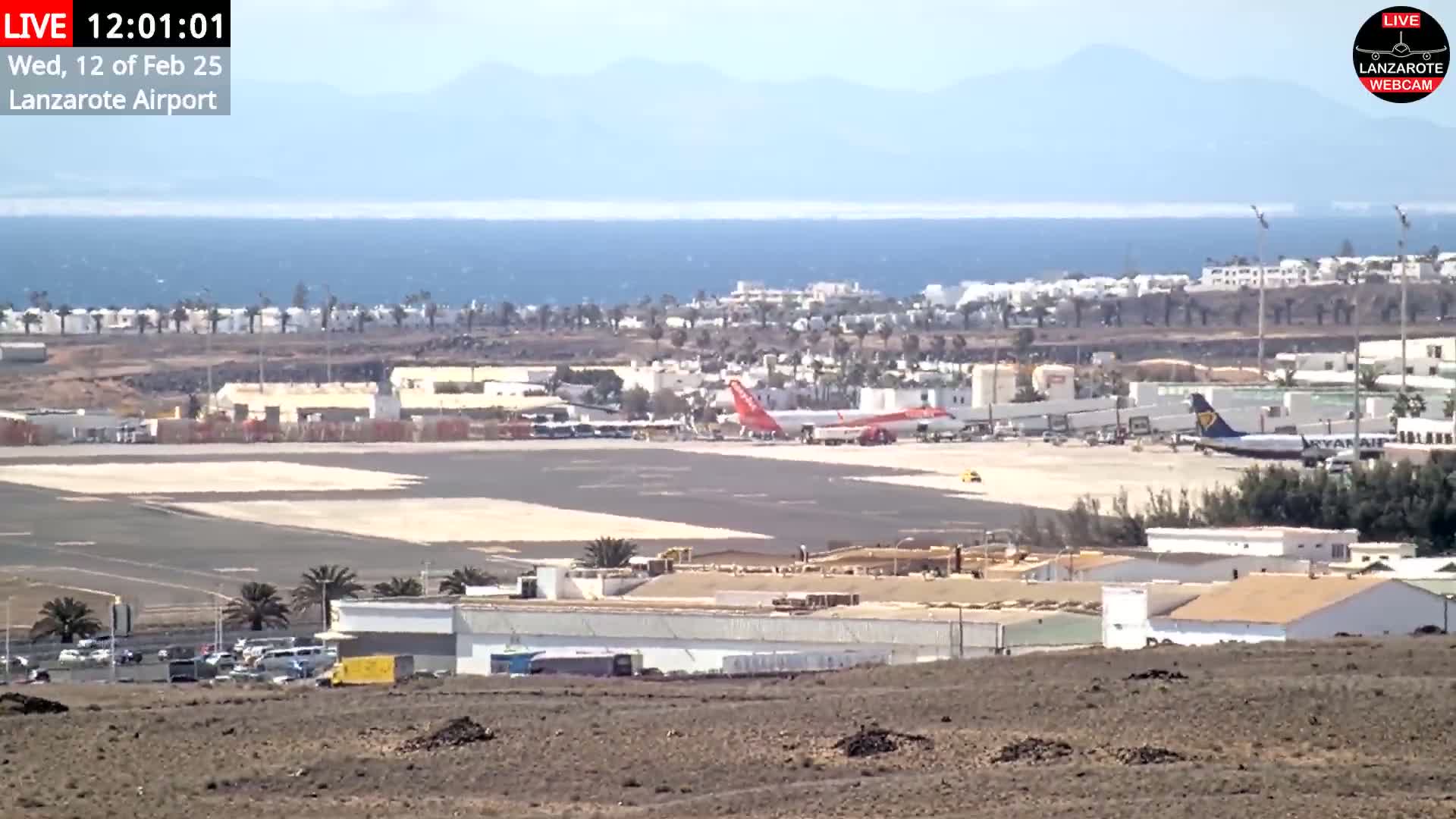 A sunny airport tarmac with two airplanes, surrounded by buildings and a body of water in the background.