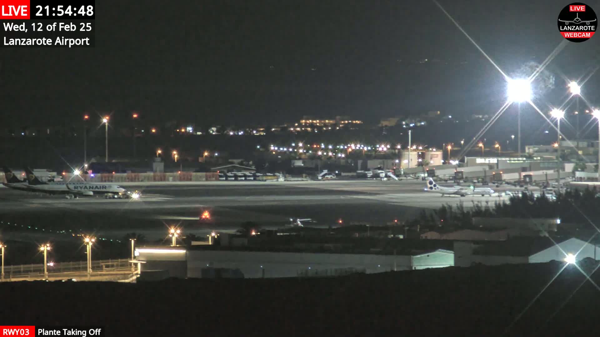 A nighttime view of an airport tarmac with several airplanes, including a Ryanair plane, under bright lights.