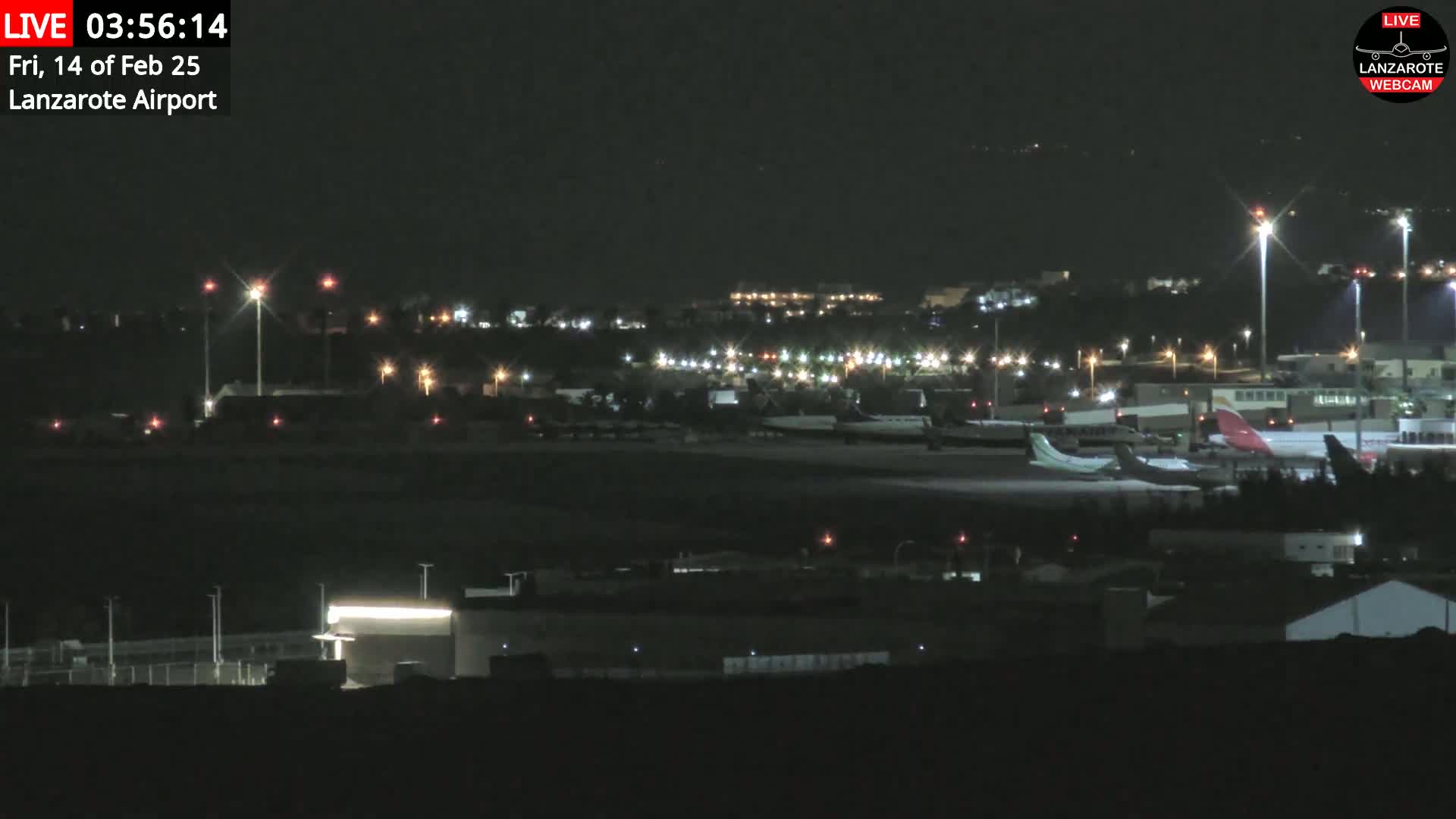 At night, several airplanes are parked on a dark airport tarmac illuminated by lights, with a city in the background.