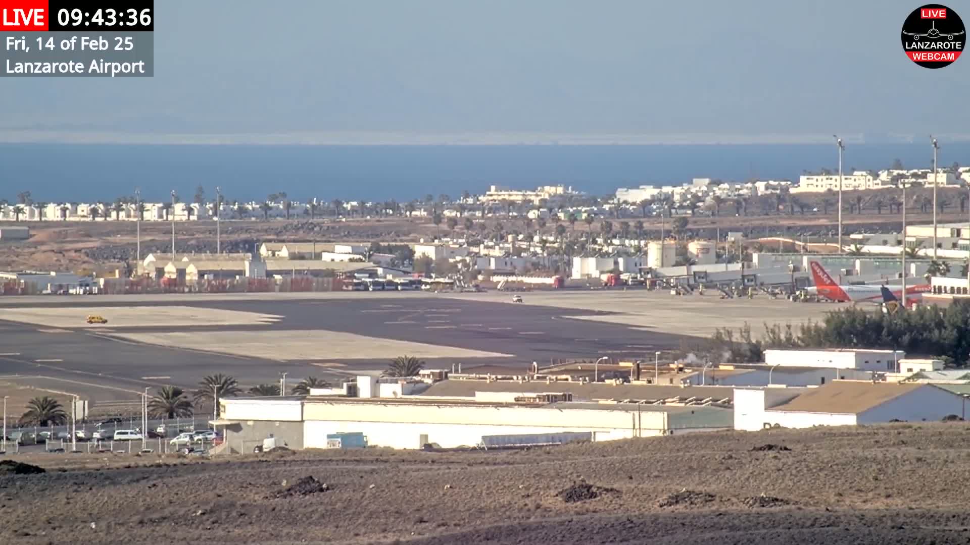A sunny day at an airport with a plane parked near a terminal, buildings, and a body of water in the background.