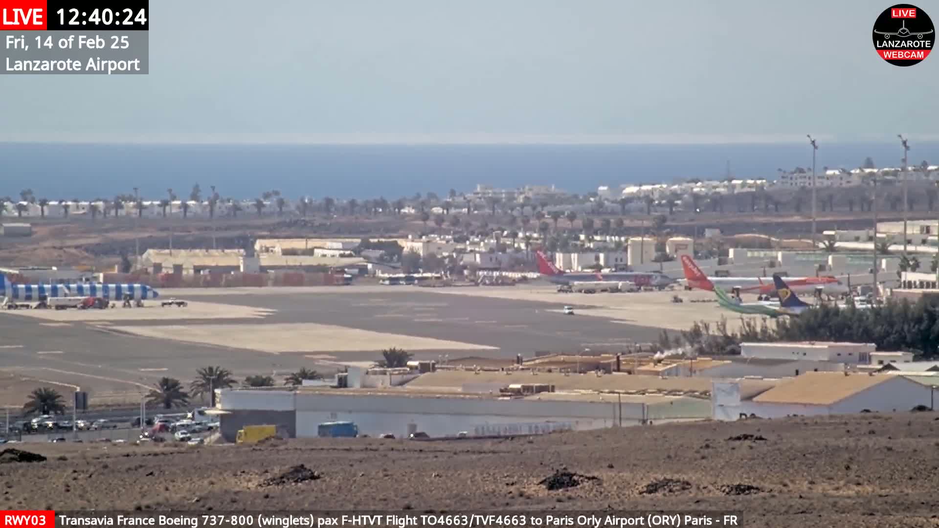 A sunny day at an airport shows several airplanes on the tarmac, with the ocean and a town visible in the background.