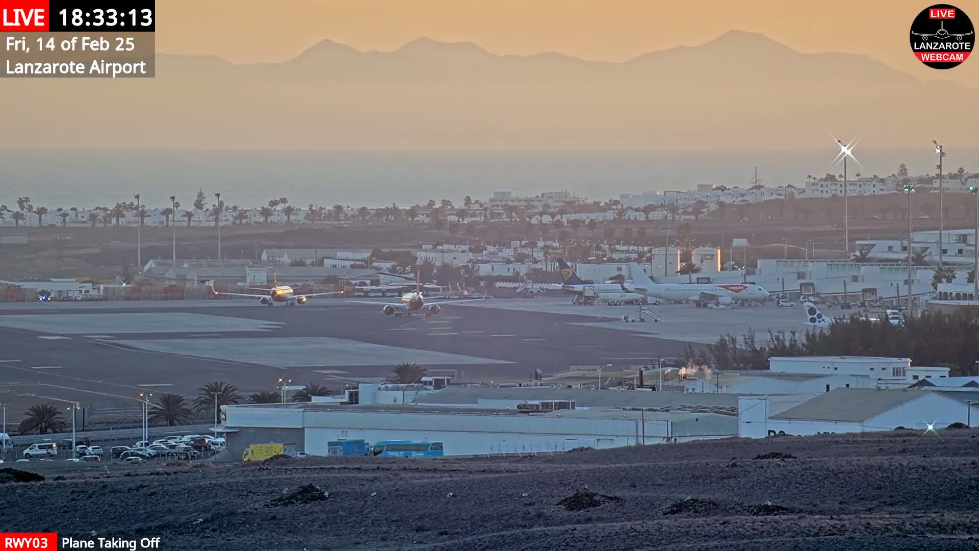 An airplane takes off from an airport runway at sunset, with mountains visible in the distance under hazy skies.