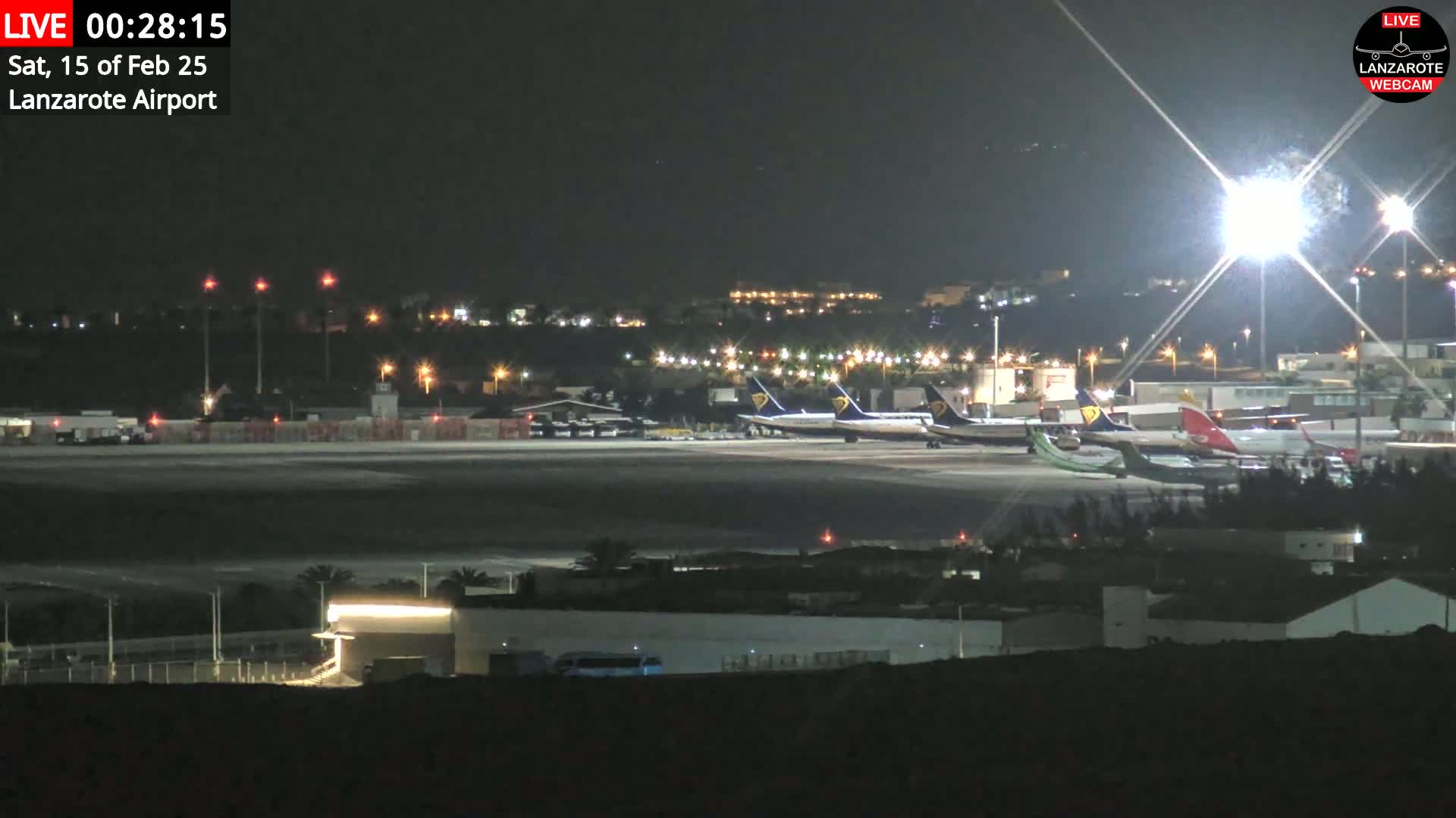 A nighttime view of a brightly lit airport tarmac with several airplanes parked, under a clear sky.