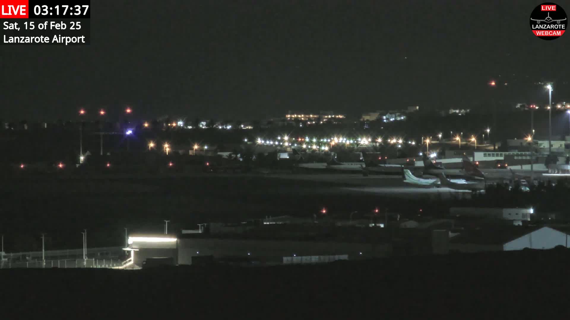 A nighttime view of an airport tarmac with several airplanes parked, surrounded by dimly lit buildings and grounds under a clear, dark sky.