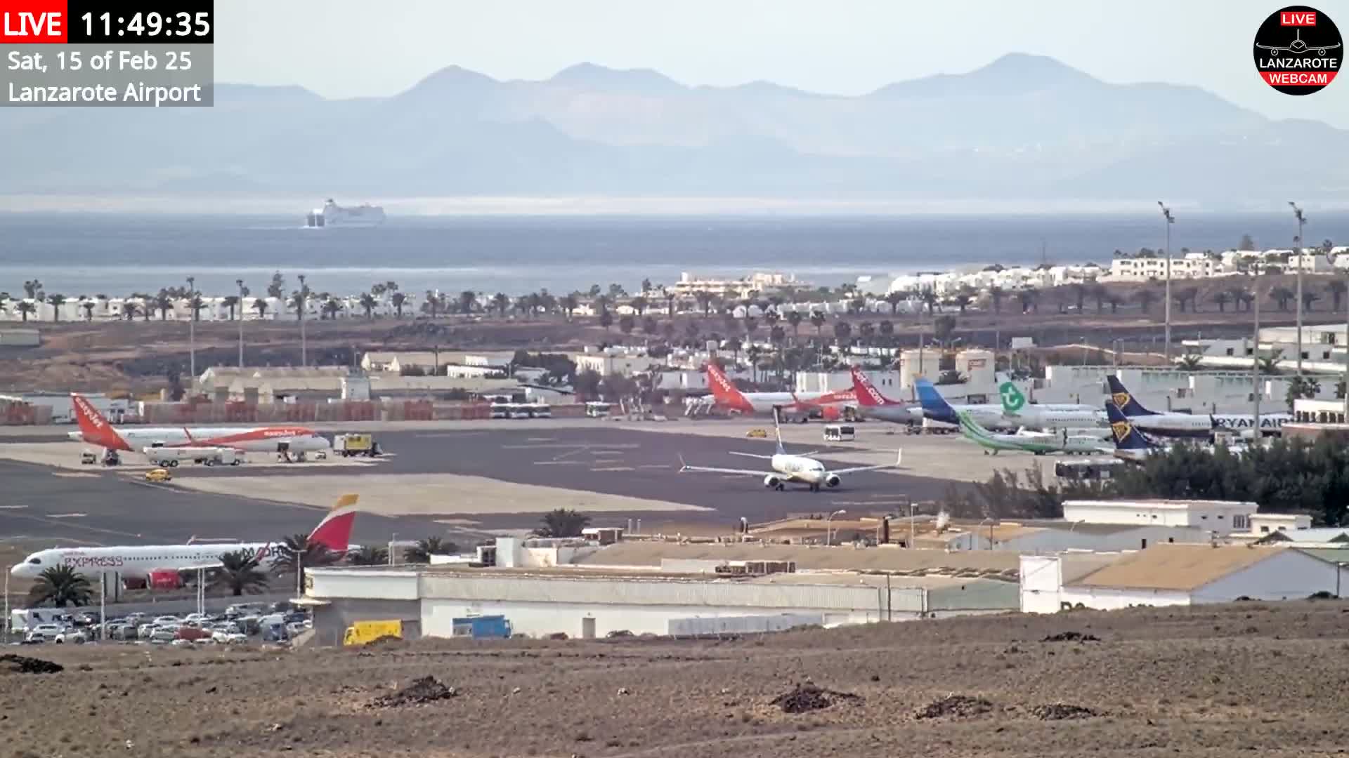 A high-angle, long shot of an airport with several airplanes on the tarmac, under hazy, sunny conditions, and a cruise ship and mountains visible in the distance.