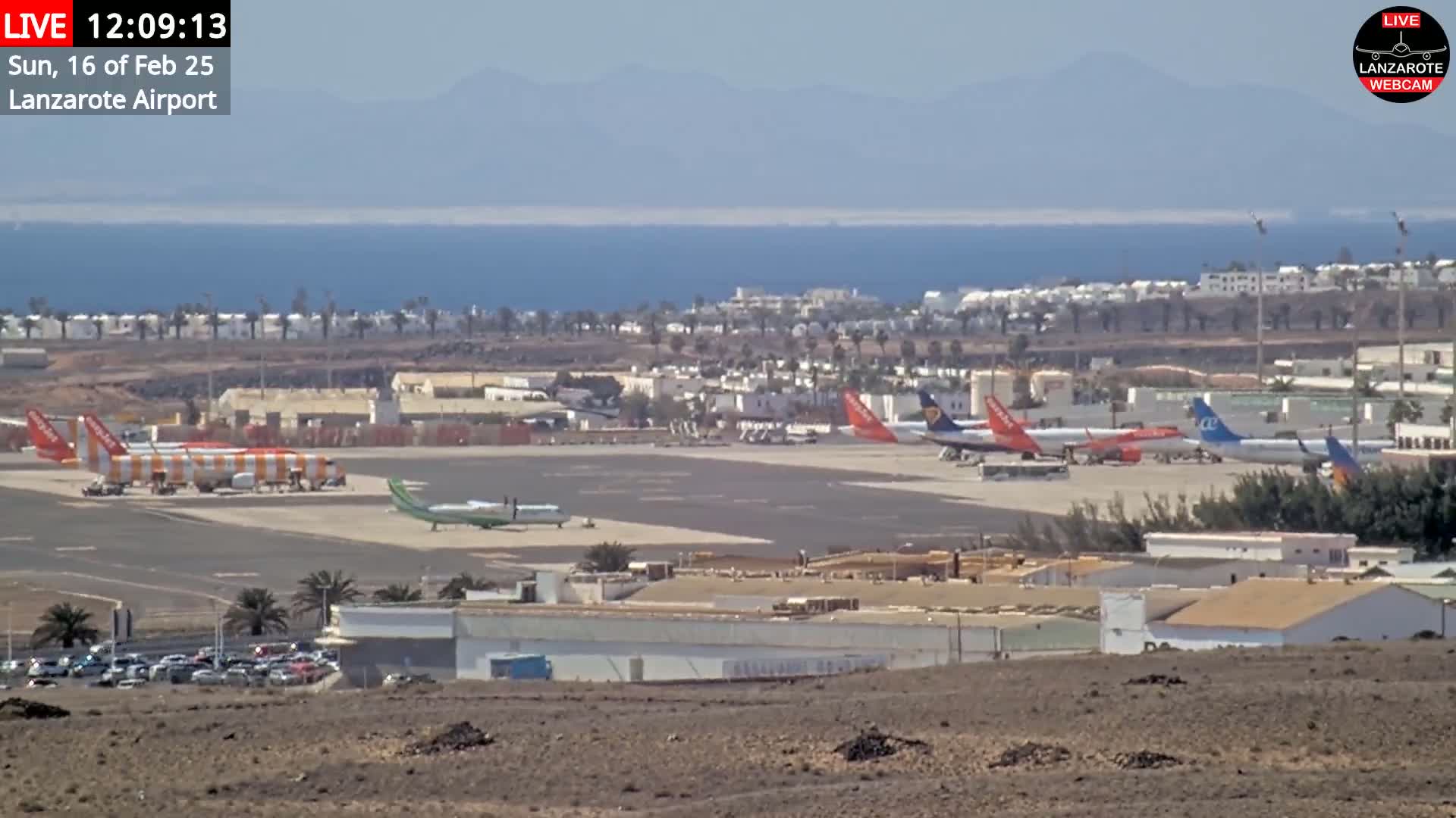 A sunny day at an airport shows several airplanes parked on the tarmac, with a body of water and distant mountains visible in the background.