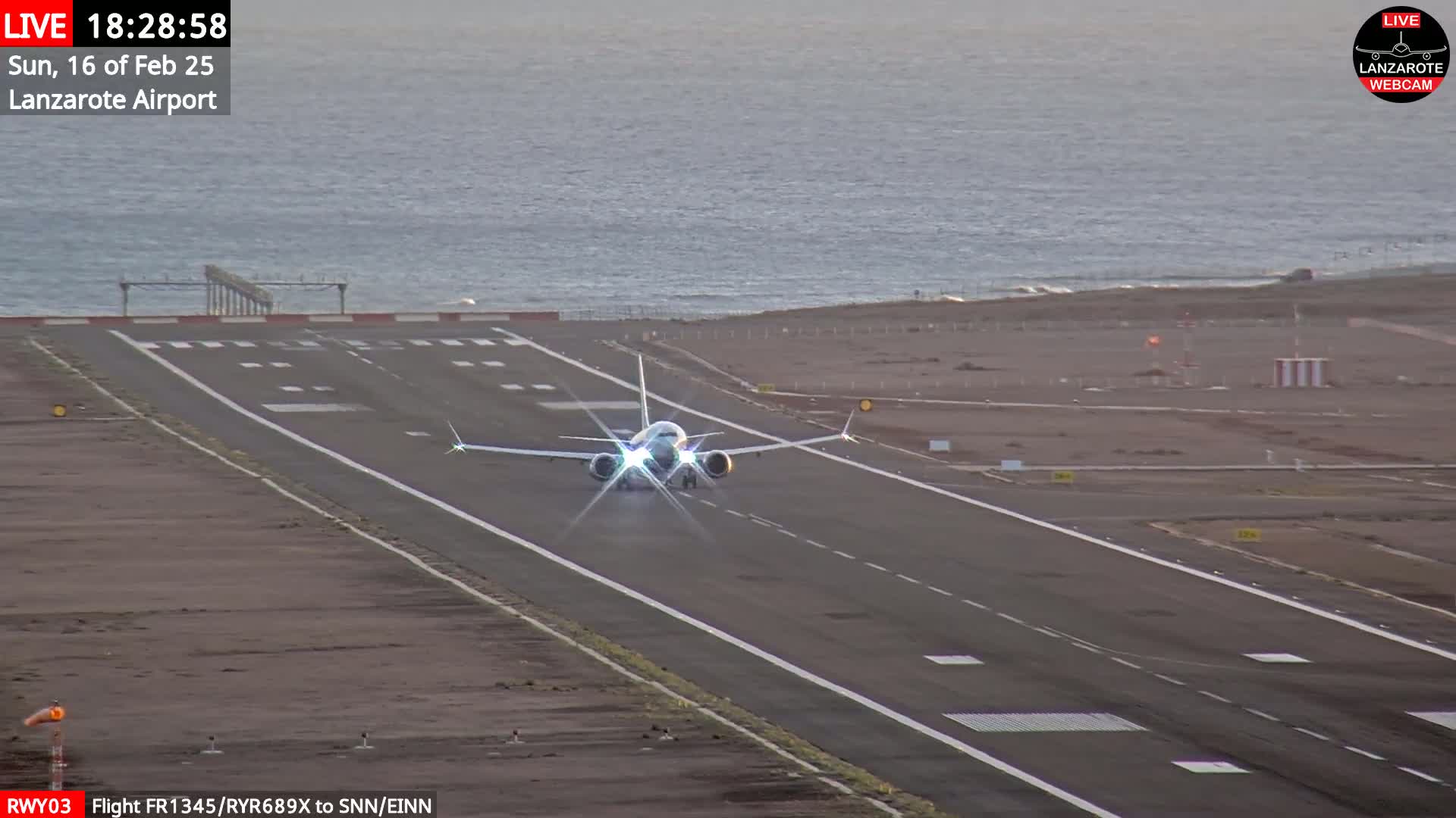 A large passenger airplane is taxiing down a runway toward the ocean under mostly clear evening skies.