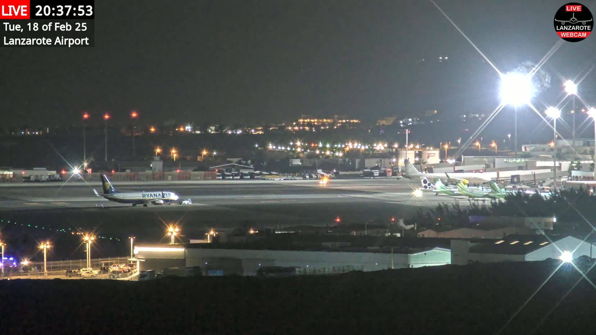 At night, a Ryanair plane sits on a runway at an airport with several other planes nearby and bright lights illuminating the tarmac.