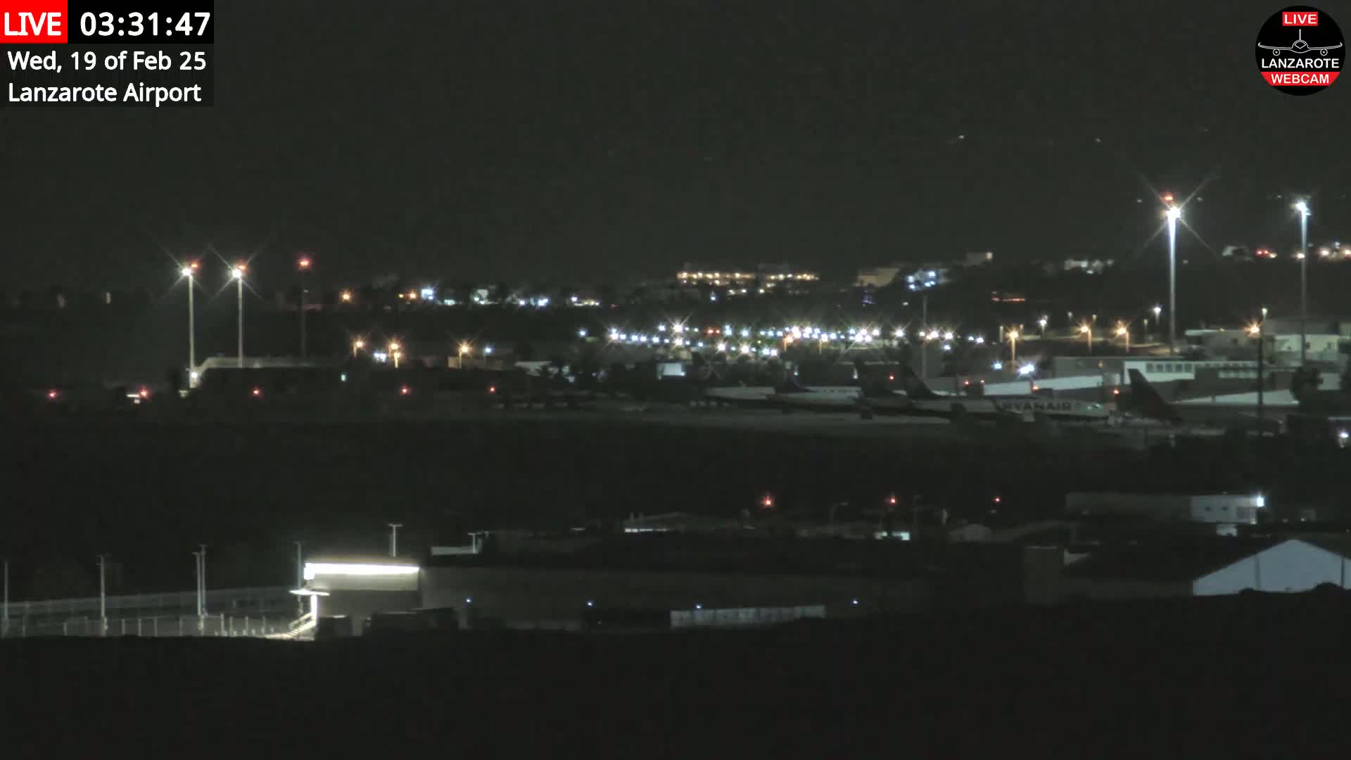 A nighttime long shot of an airport tarmac with several airplanes parked and illuminated buildings.
