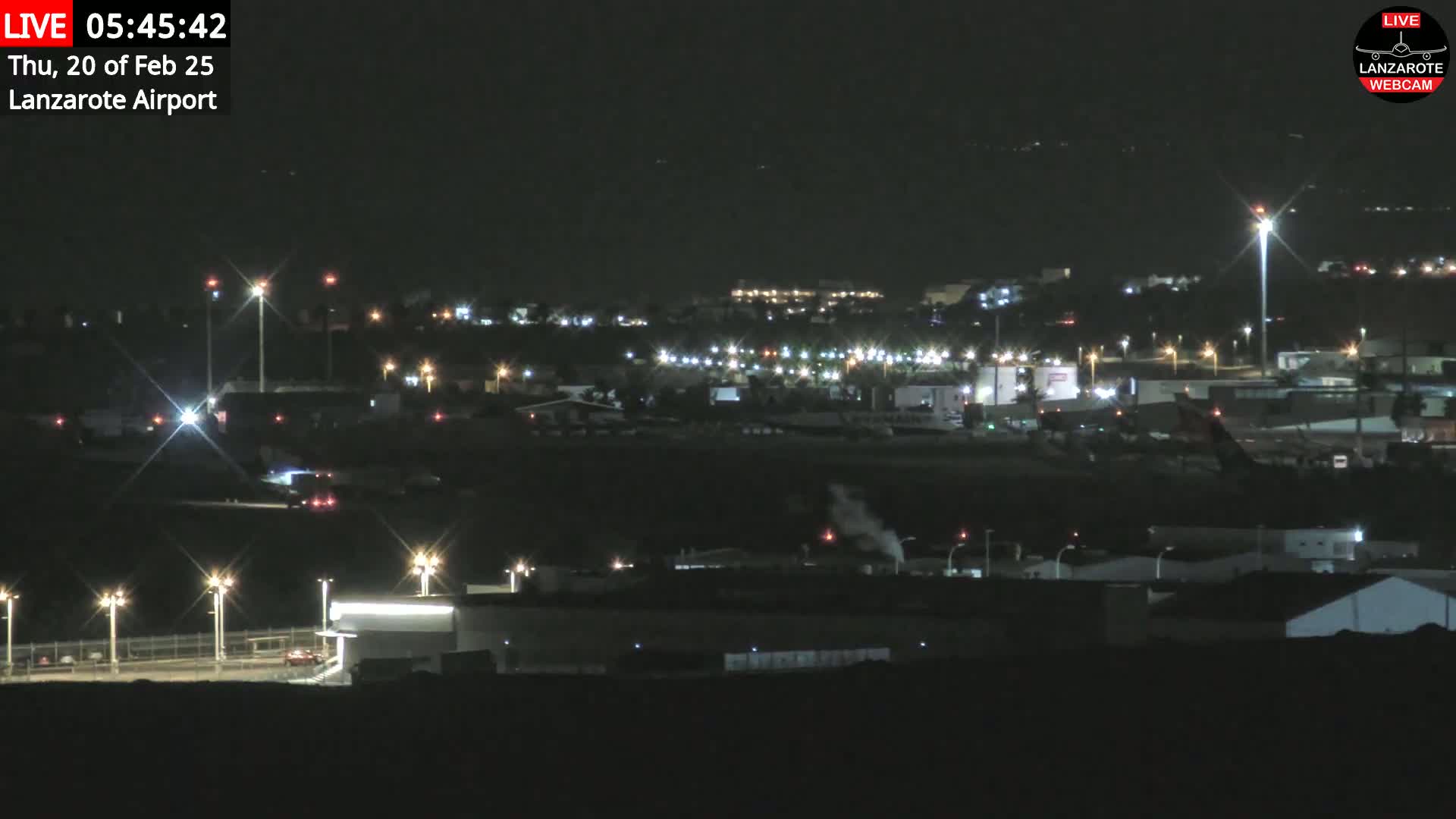 A nighttime long shot of an airport at night, illuminated by lights, with a hazy or smoky atmosphere.