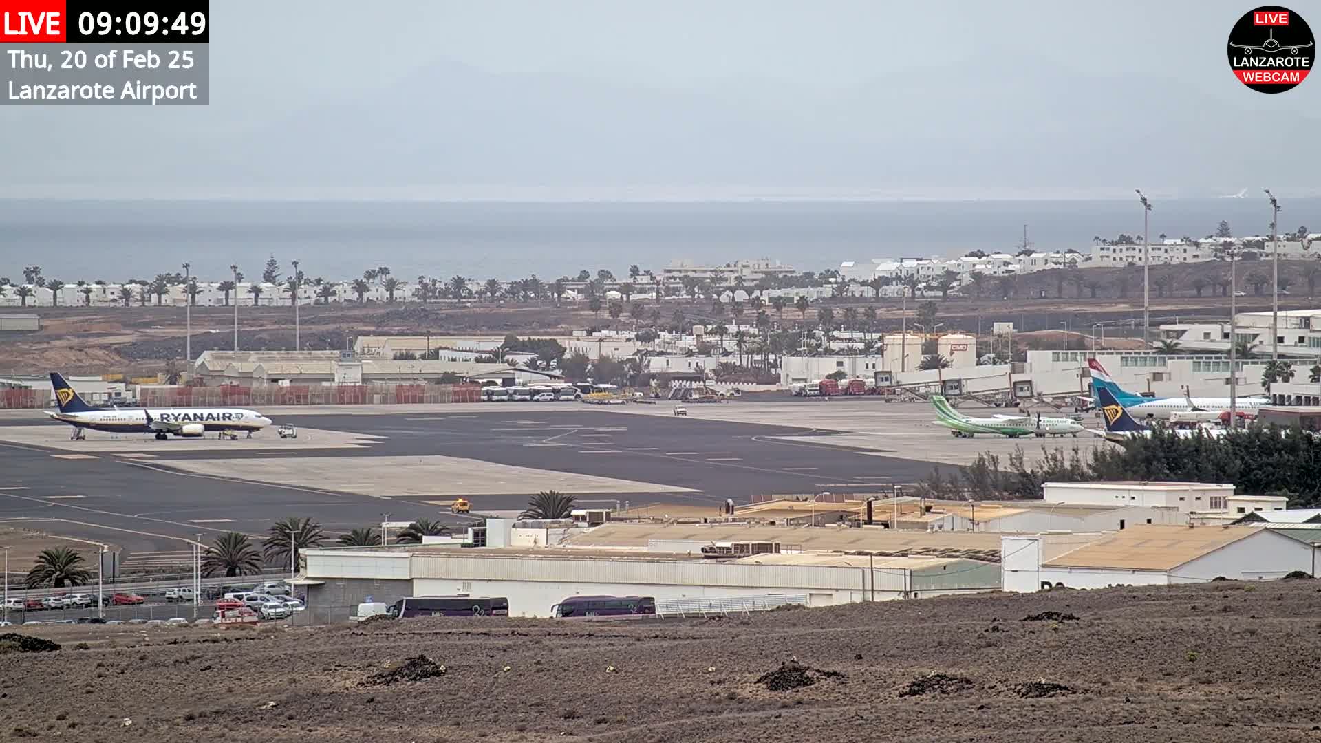 An airport tarmac with several airplanes, airport buildings, and a distant ocean under an overcast sky.