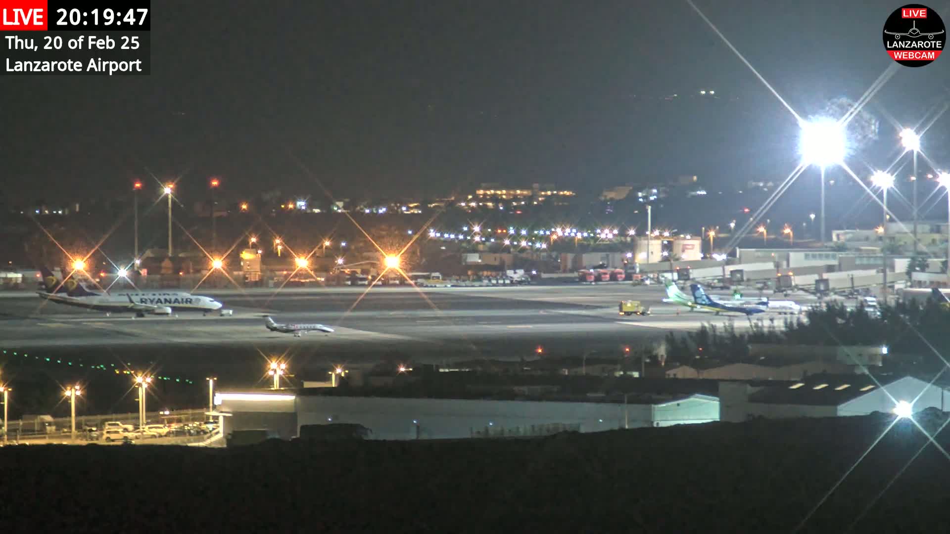 A nighttime view of a brightly lit airport tarmac with several airplanes parked and one taxiing, under a clear, dark sky.