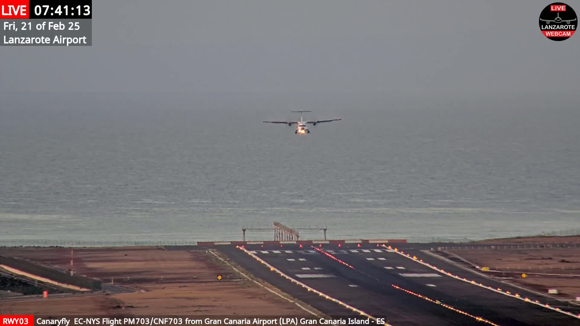 A turboprop airplane is making a low approach to land on a runway that borders the ocean under overcast skies.