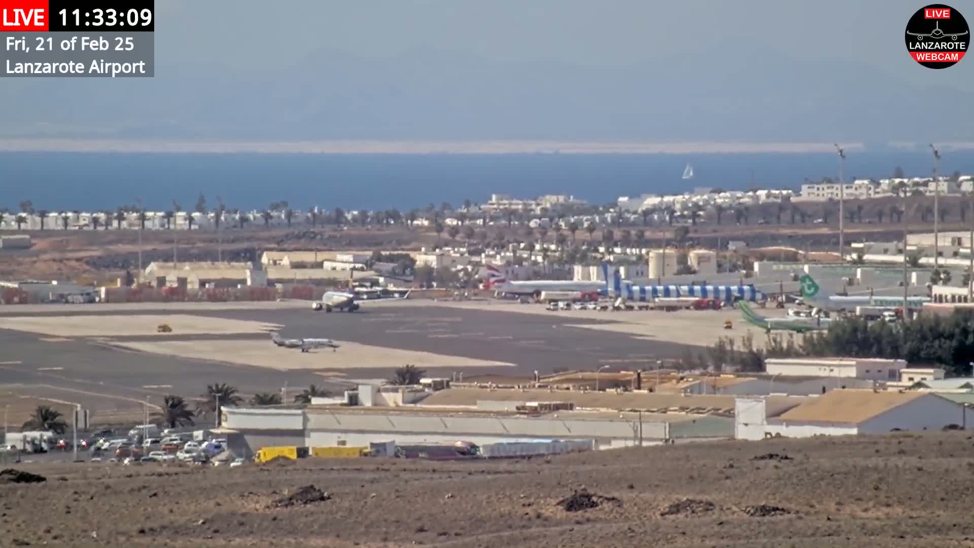 A high-angle, long shot of an airport tarmac with several planes, airport buildings, and vehicles, overlooking a calm ocean under a clear sky.