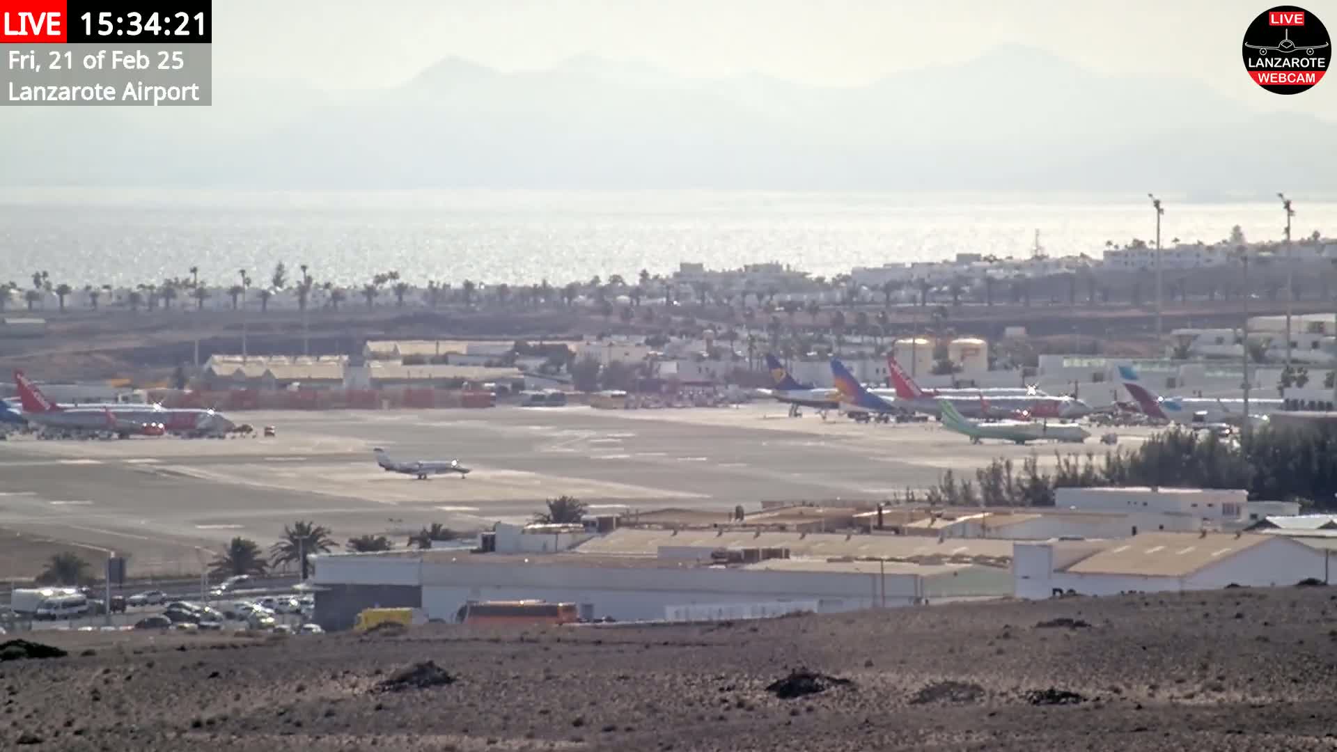 A hazy, sunny day shows a view of an airport tarmac with several airplanes, airport buildings, and vehicles, beyond which lies a body of water and distant mountains.