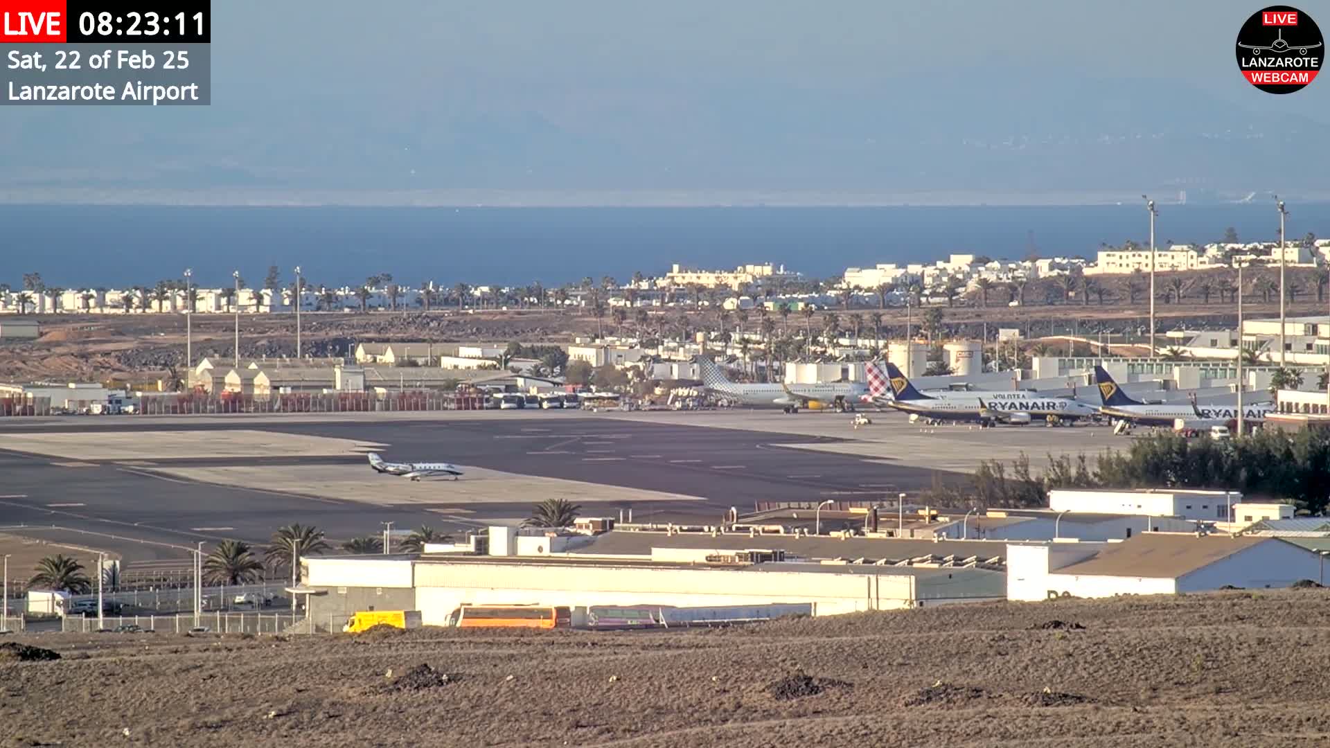 A high-angle, long shot of an airport tarmac with several airplanes, airport buildings, and palm trees, overlooking a calm ocean under a clear sky.