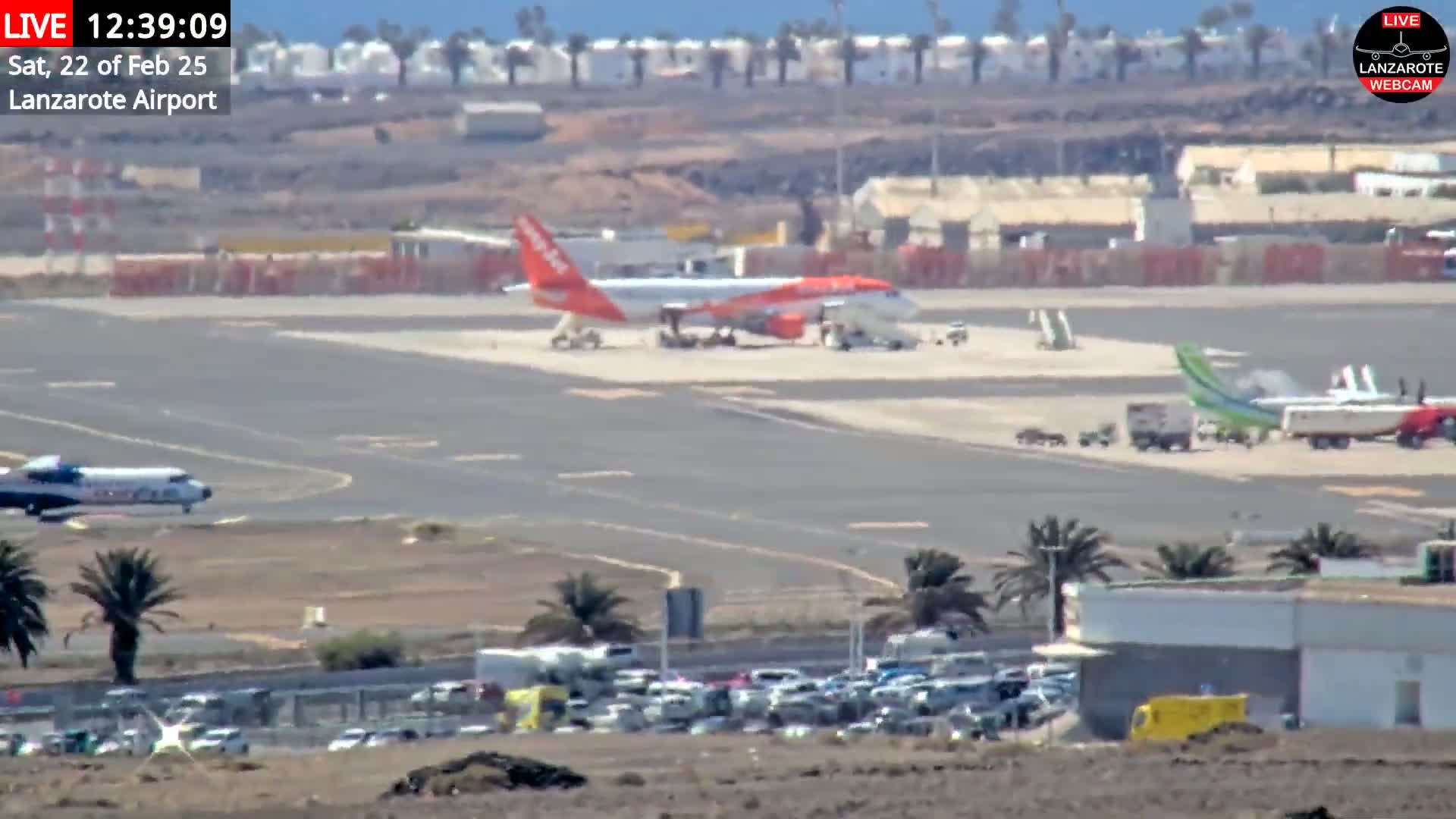 An airport tarmac on a sunny day shows several airplanes, ground support vehicles, and a large parking lot full of cars.