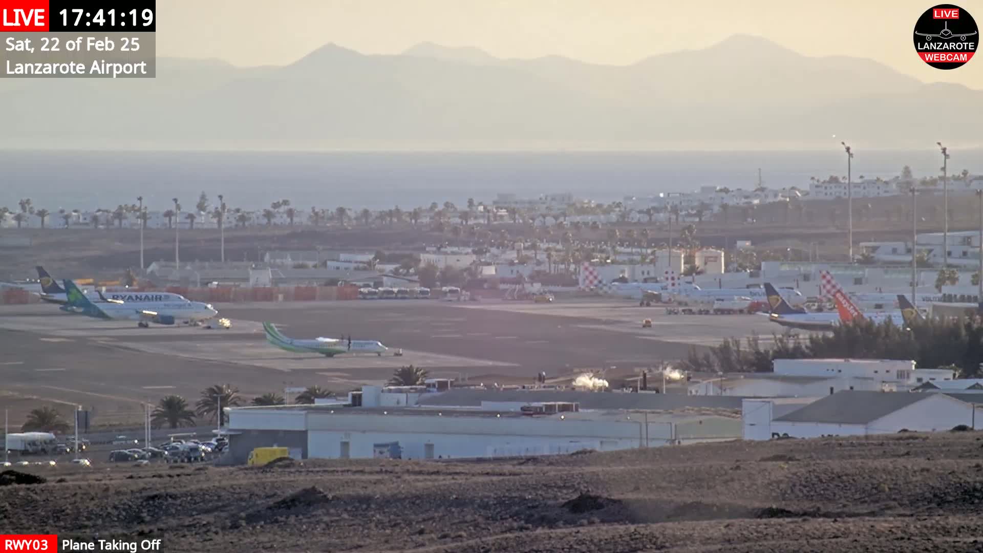 An airport tarmac with several airplanes, including a Ryanair plane, is visible against a hazy background of a coastline and distant mountains under a mostly clear sky.