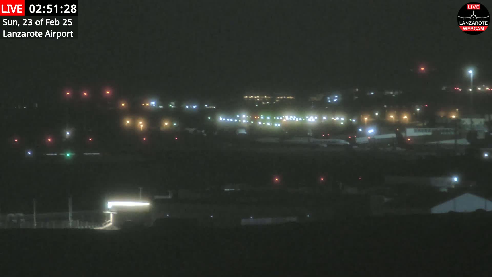 A nighttime view of an airport with numerous lights illuminating the tarmac and surrounding buildings, under hazy or foggy conditions.