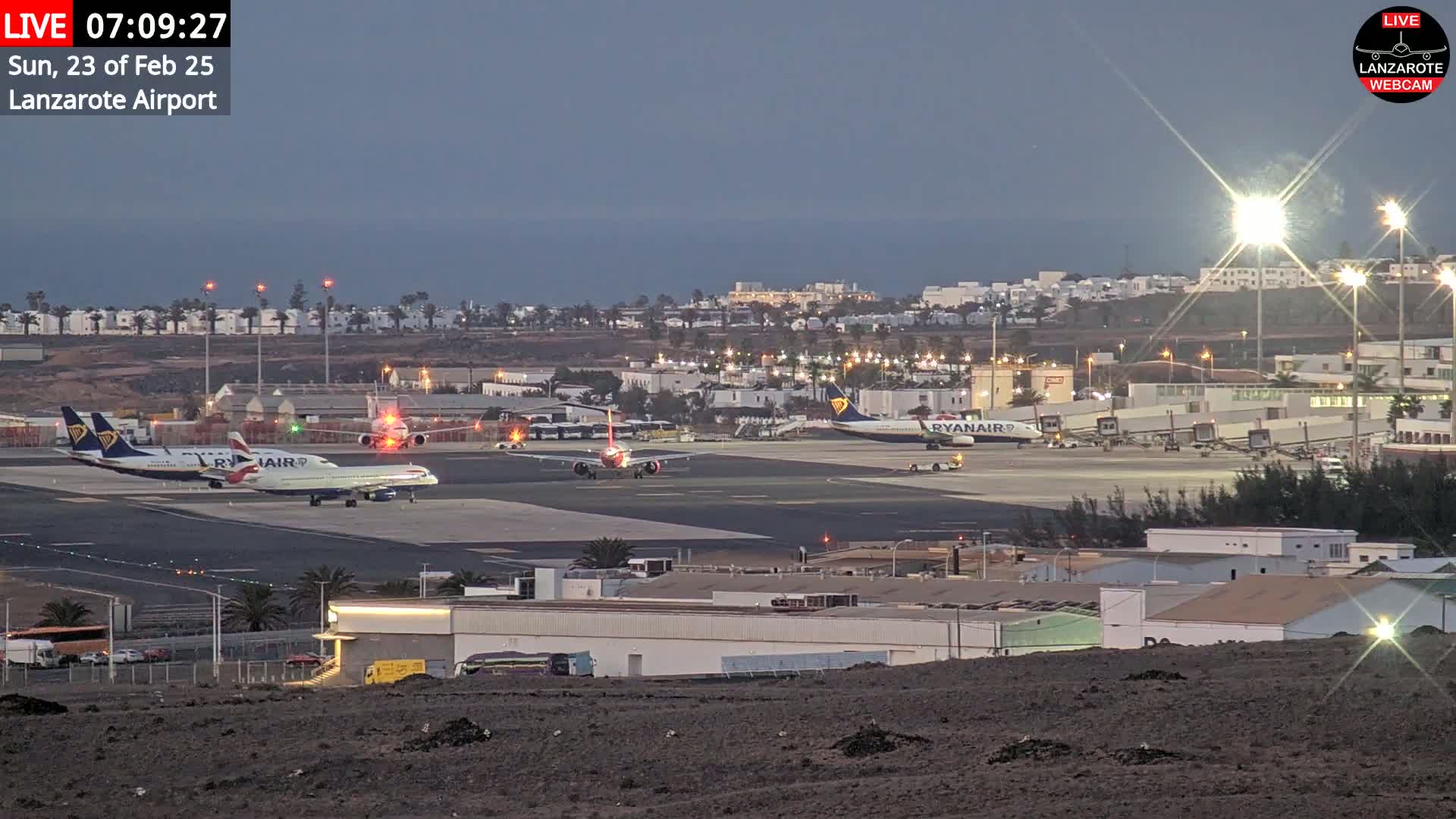 Several airplanes are parked on an airport tarmac at twilight, with a body of water visible in the distance.