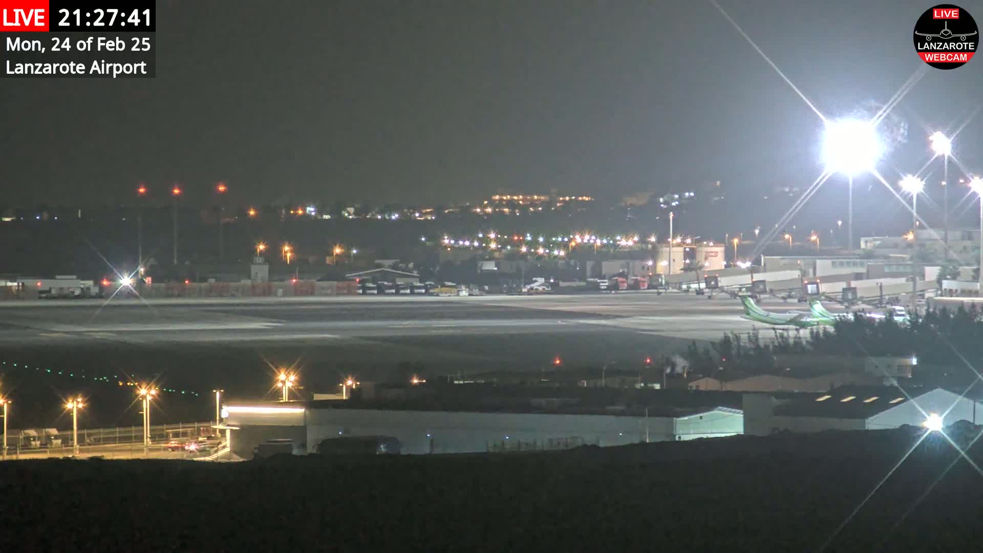 A brightly lit airport tarmac at night shows an airplane and various airport vehicles, with a darker, hilly landscape visible in the background under a clear, dark sky.