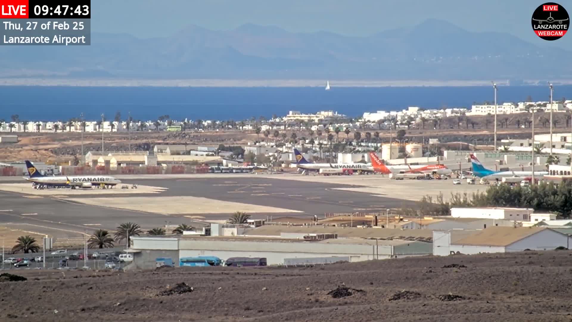 Under a clear sky, several airplanes are parked on an airport tarmac near a body of water and a town.