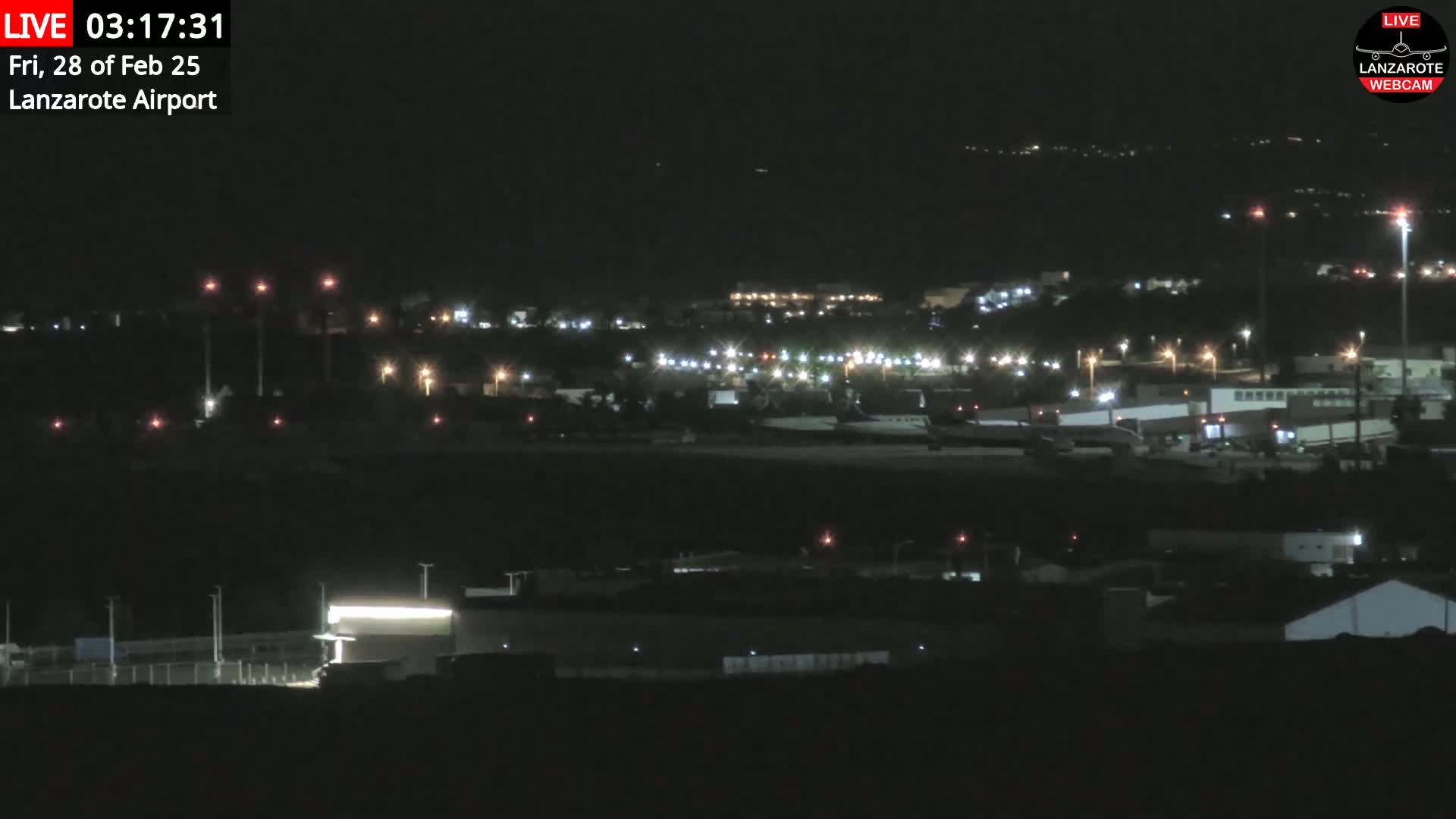 A nighttime long shot of an airport with scattered lights illuminating the tarmac and surrounding buildings under a clear, dark sky.