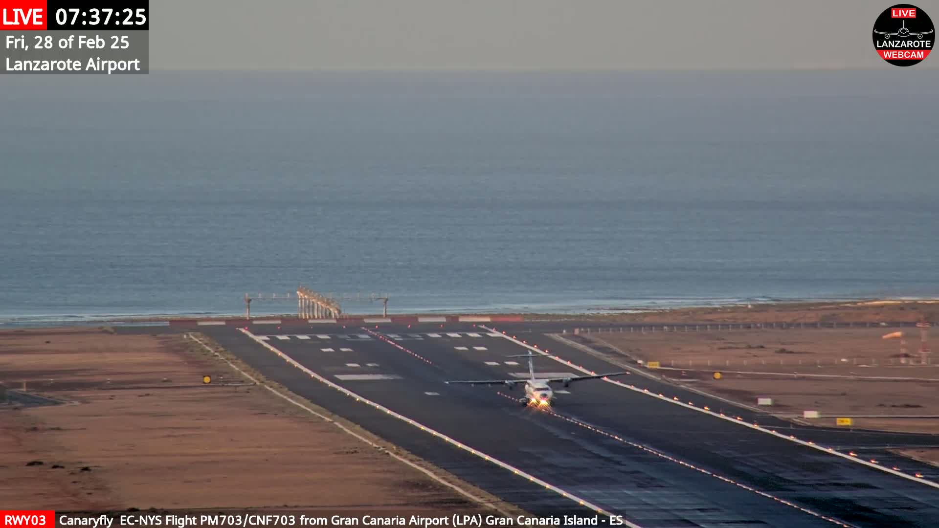A small airplane taxis on a runway next to a calm ocean under a mostly clear sky.