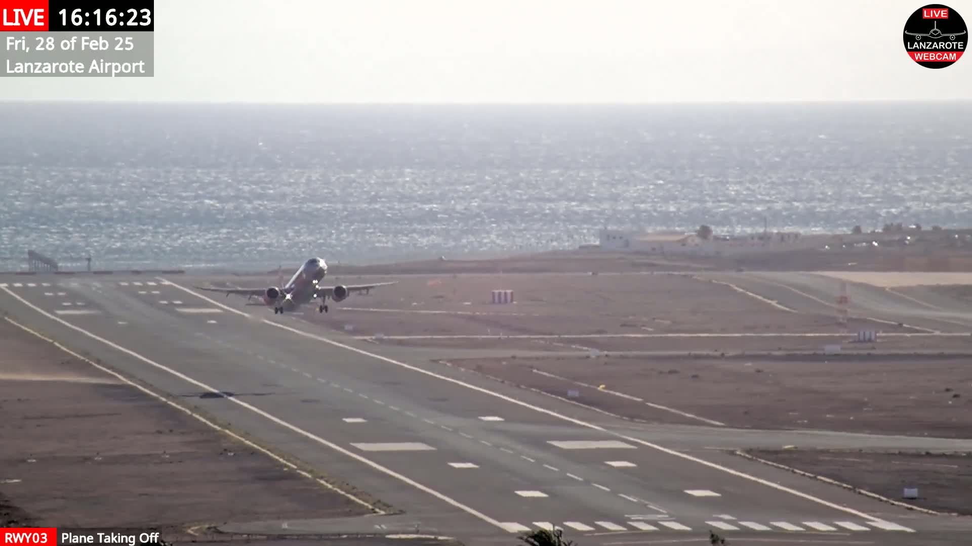 A plane takes off from a runway near a body of water under bright, sunny conditions.