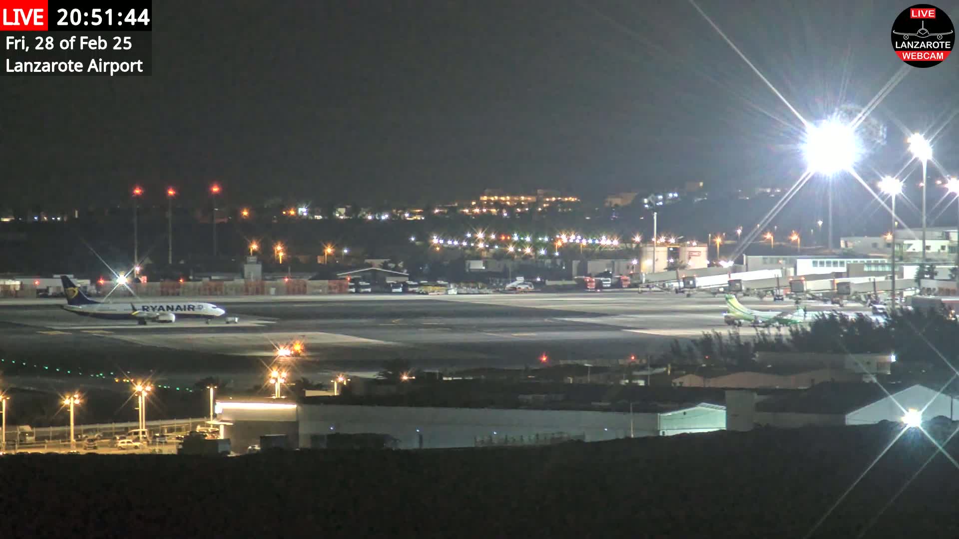 A nighttime view of an airport tarmac with several airplanes, buildings, and bright lights illuminating the scene.