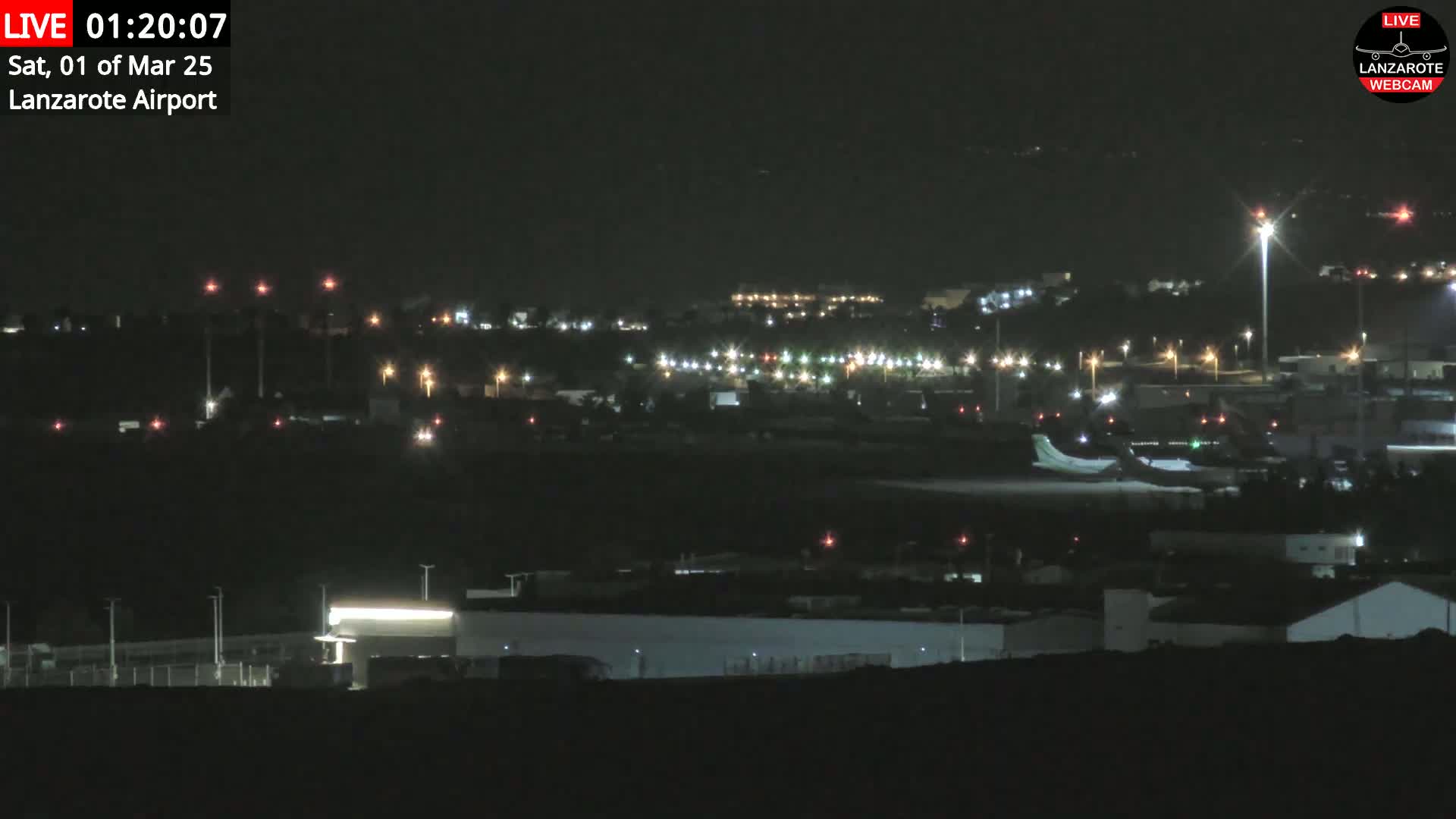 A nighttime, long shot of an airport with a plane on the runway and surrounding buildings and lights faintly visible under a dark sky.