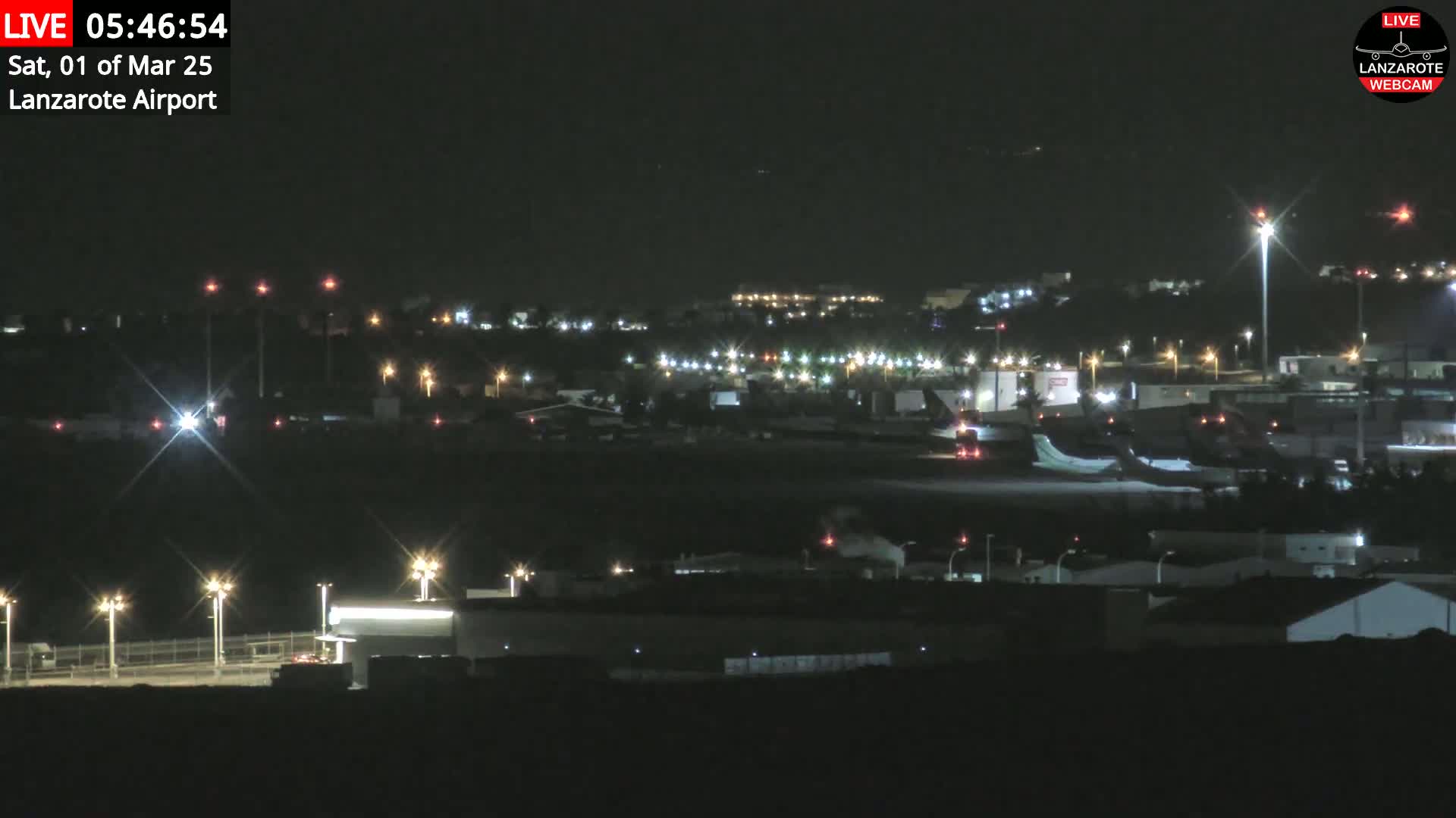 A nighttime view of an airport runway with lights illuminating various buildings and a plane on the tarmac under a clear, dark sky.