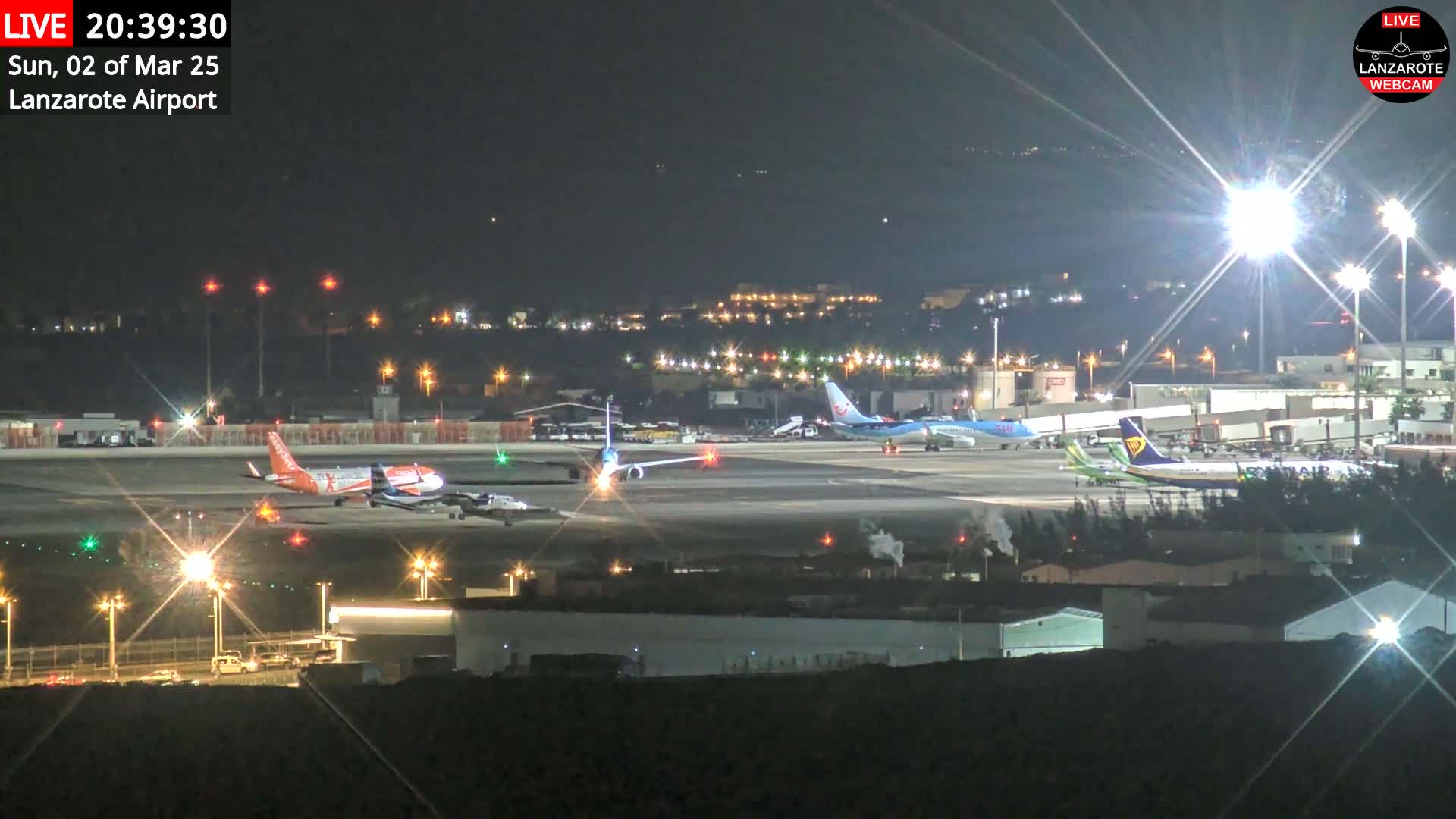 A nighttime view of an airport tarmac with several airplanes parked and one taxiing, illuminated by bright lights.