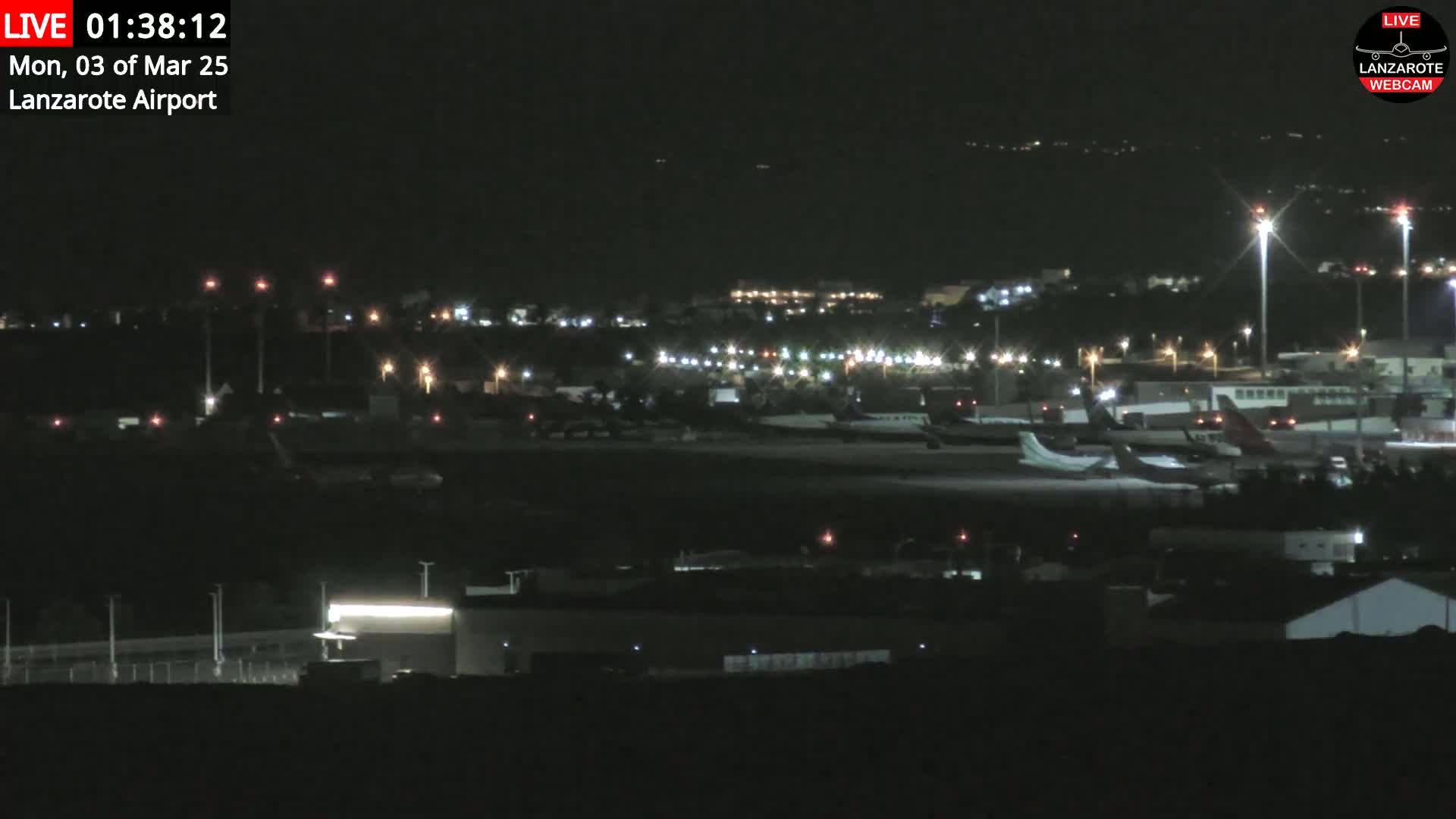 A nighttime long shot of an airport tarmac with several airplanes parked, illuminated by runway and building lights under a clear, dark sky.