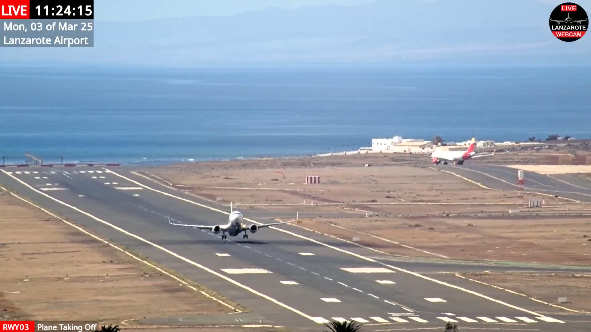 A plane takes off from an airport runway next to a calm ocean under a clear sky.